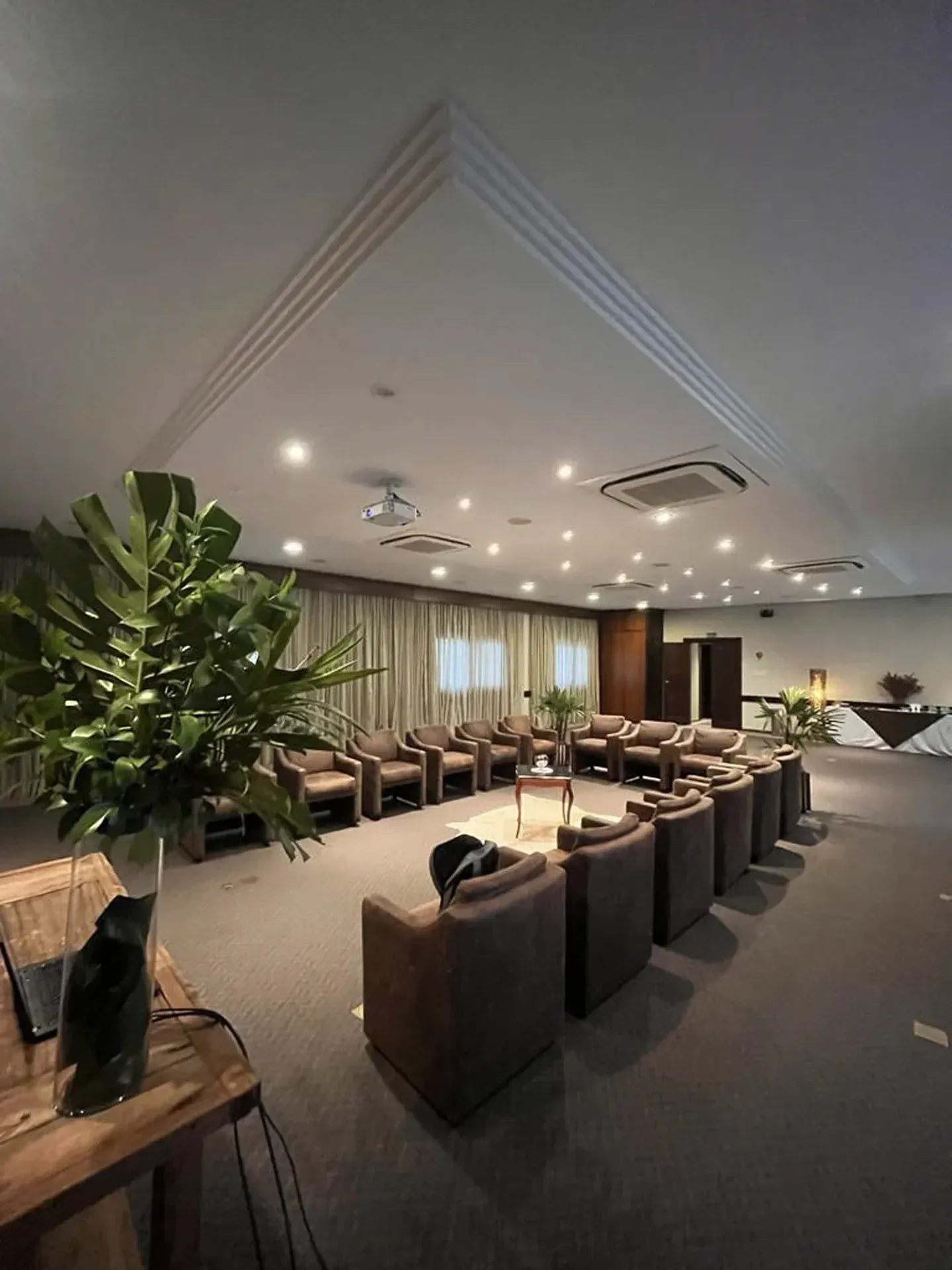 Sala de conferências moderna com cadeiras, mesas, plantas e iluminação embutida no teto.