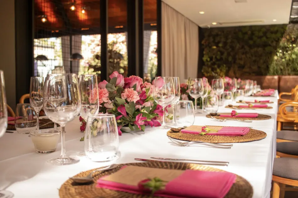 Mesa de banquete elegantemente posta com arranjos de flores rosa, taças de vinho e detalhes em dourado nos lugares marcados.