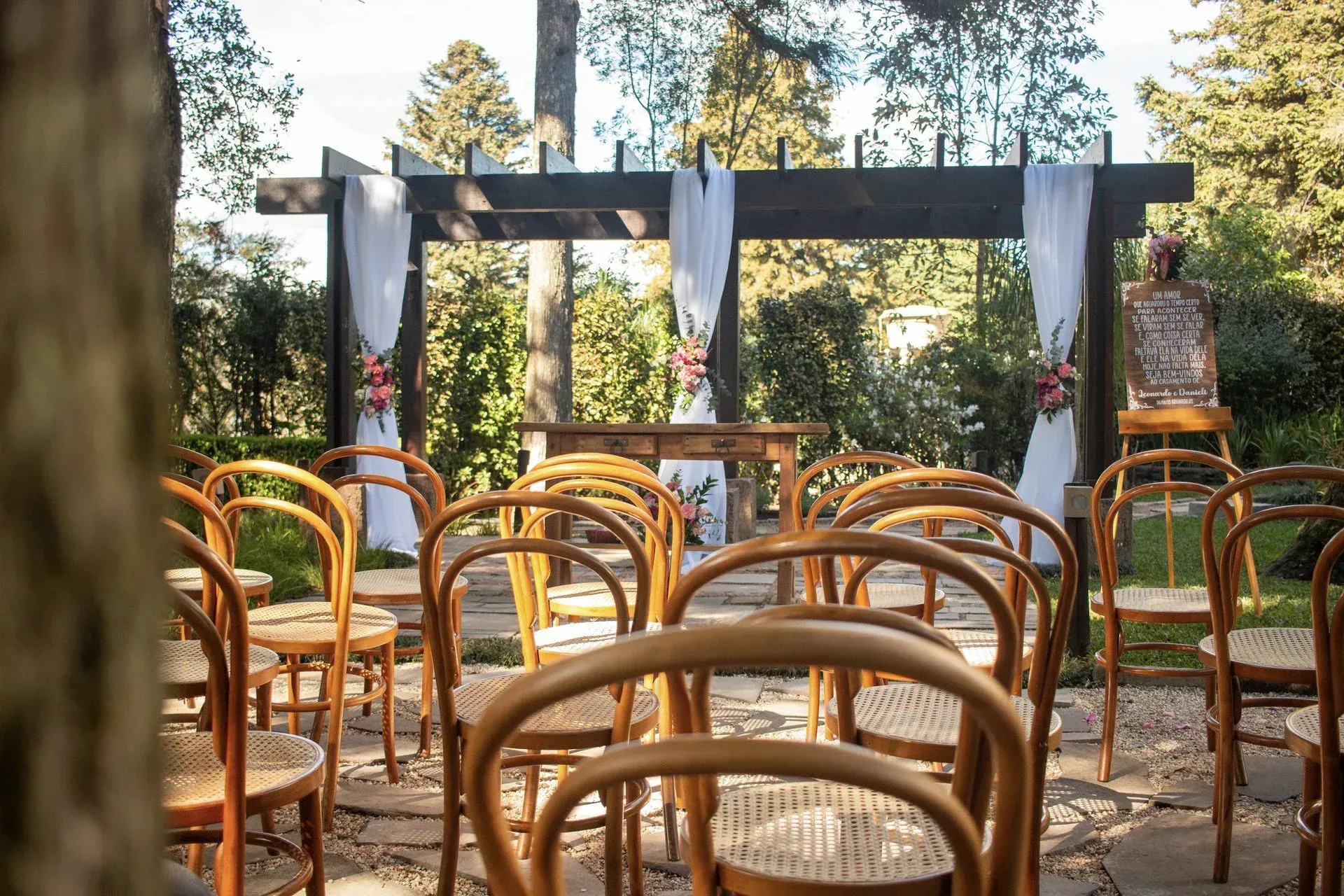 Cerimônia ao ar livre com cadeiras de madeira e pérgola com cortinas azuis em um cenário de floresta.