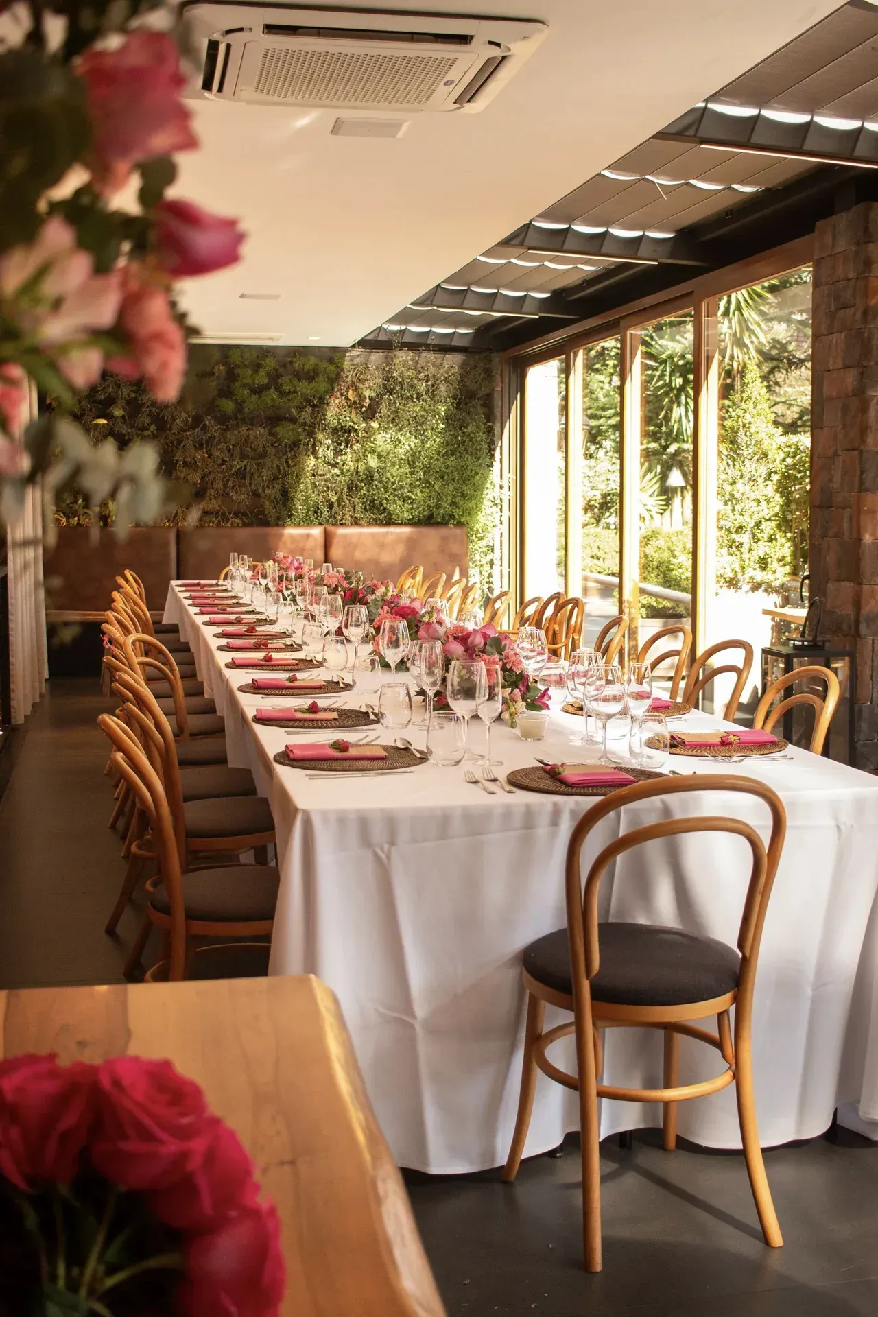 Elegante mesa comprida para banquetes, posta com toalhas brancas e flores, ao lado de grandes janelas em uma sala de jantar ensolarada.