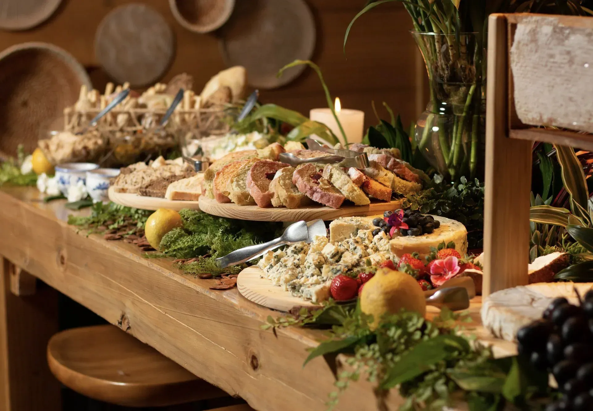 Mesa de buffet rústica com travessas variadas de comida, pão, verduras e folhagens decorativas.