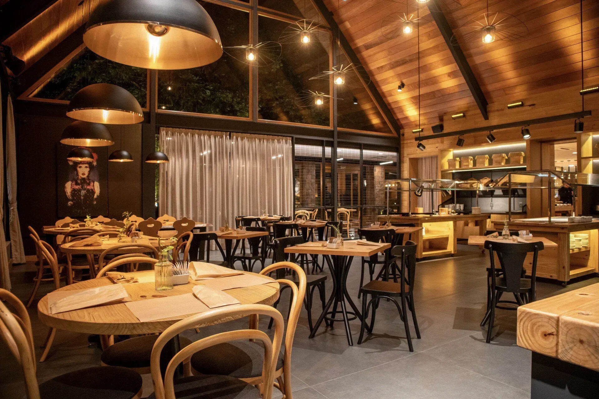 Interior aconchegante do restaurante com painéis de madeira, mesas redondas, cadeiras de vime, luminárias pendentes e uma área de bar acolhedora.