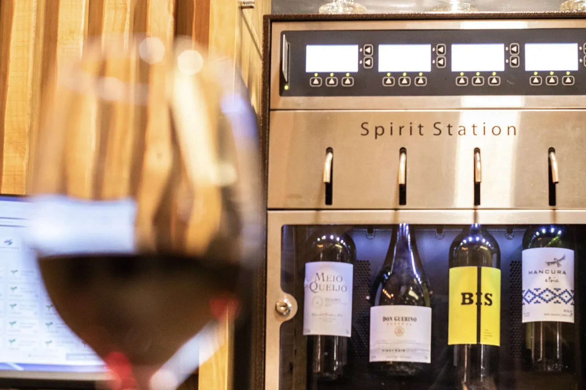 Dispensador de garrafas de vinho com a etiqueta “Spirit Station” atrás de uma taça de vinho desfocada.