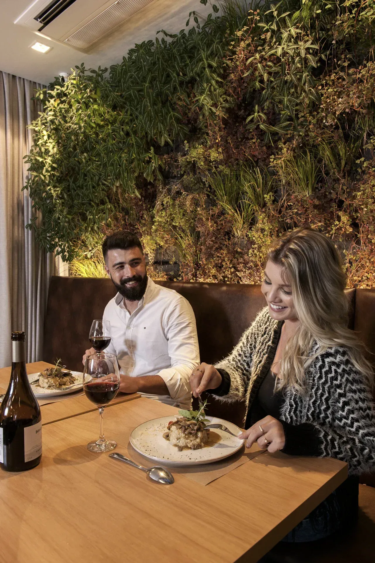 Duas pessoas jantando em uma mesa de madeira ao lado de uma exuberante parede verde em um restaurante com iluminação aconchegante.