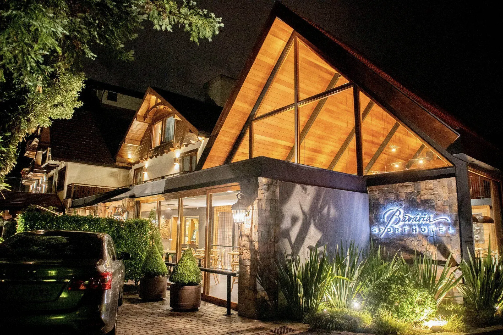 Chalé iluminado em forma de A à noite, com luzes aconchegantes, entrada de pedra e letreiro de neon perto de um carro estacionado.