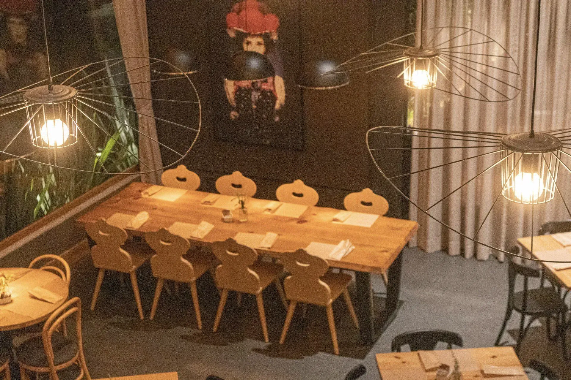 Interior aconchegante de café com mesas de madeira, bancos, luminárias pendentes em tons quentes e um mural decorativo na parede.