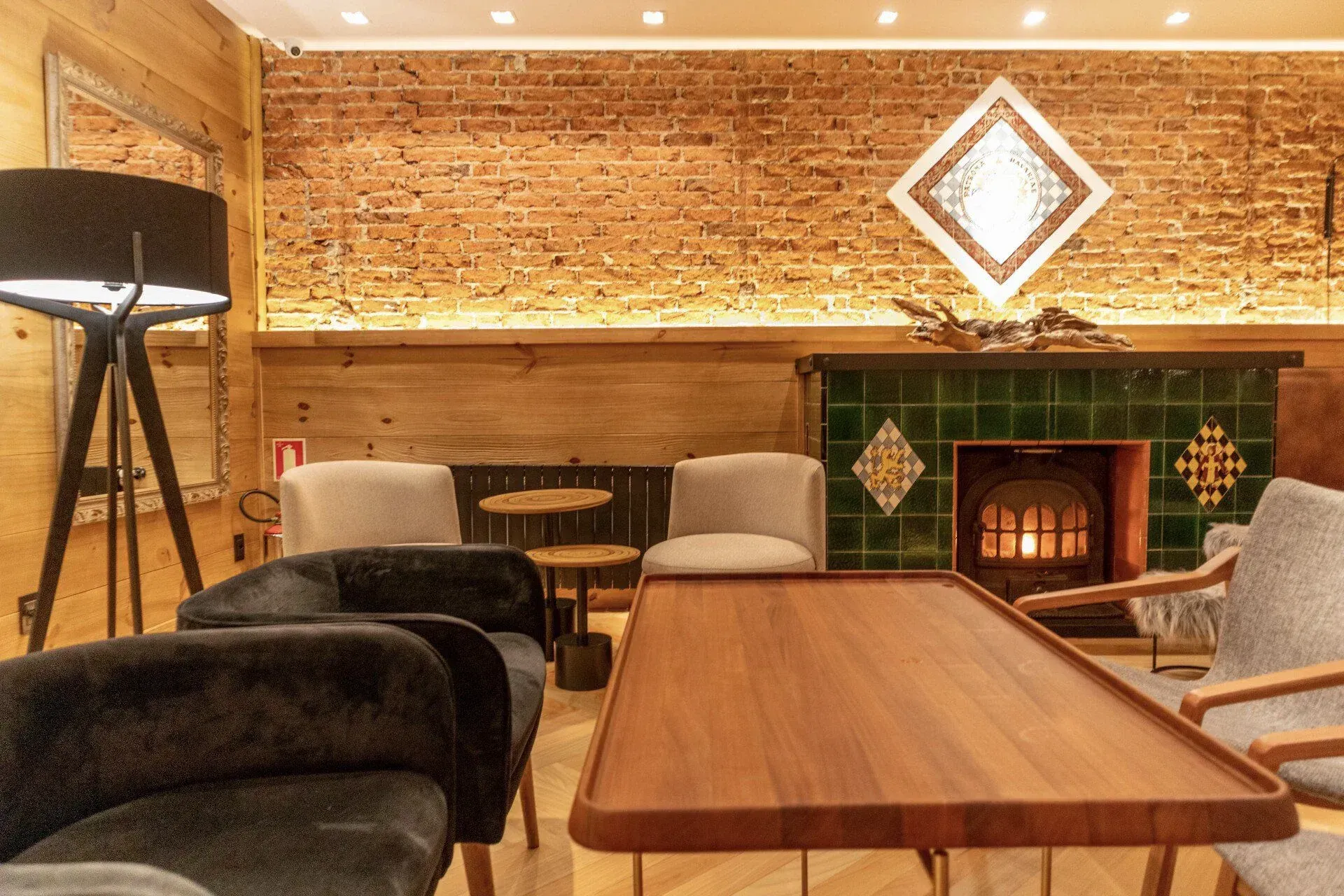 Sala de estar aconchegante com parede de tijolos, lareira, mesas de madeira e cadeiras almofadadas sob iluminação quente.