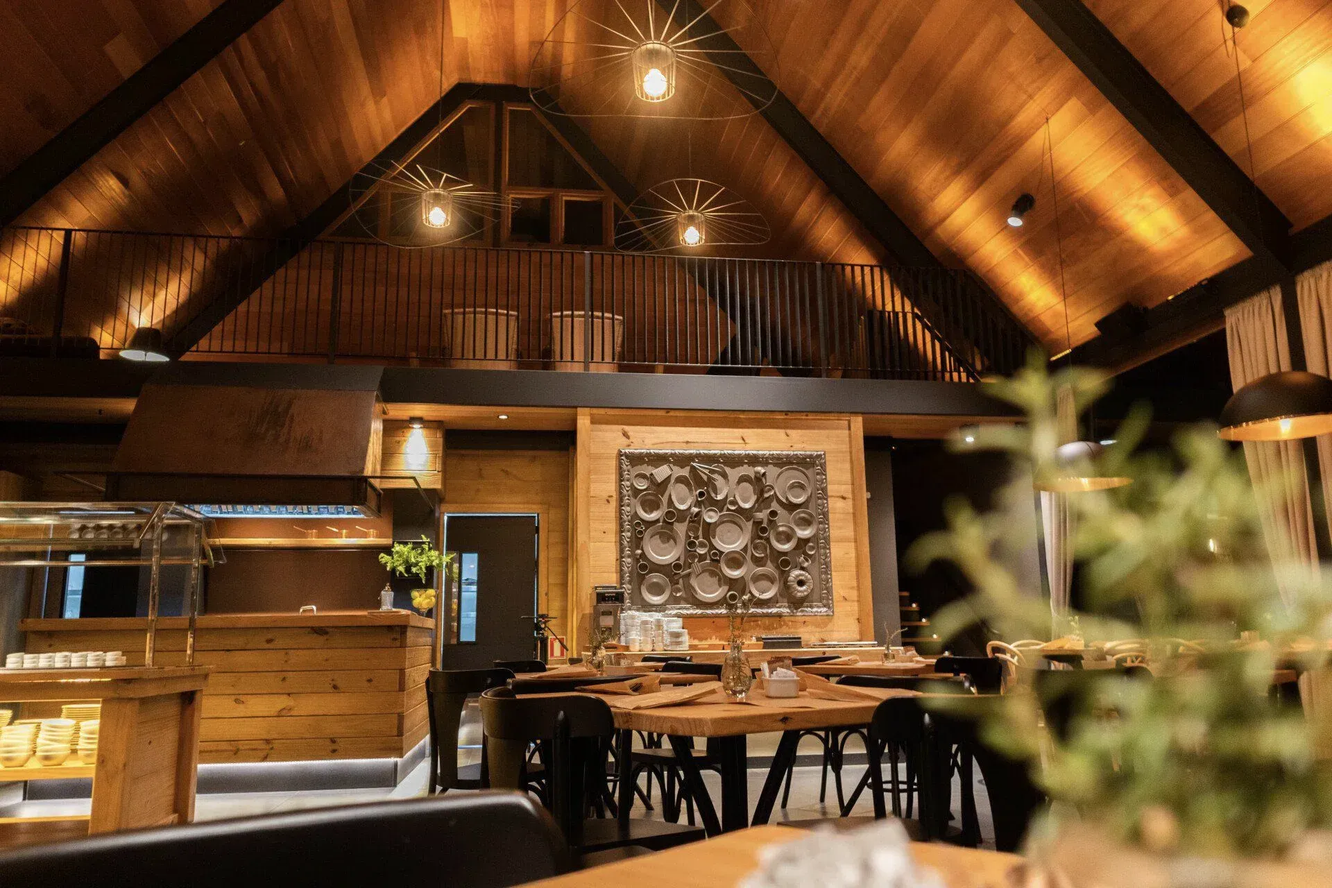 Interior aconchegante de restaurante em madeira com teto alto em abóbada, luminárias pendentes e mesas cuidadosamente arrumadas.