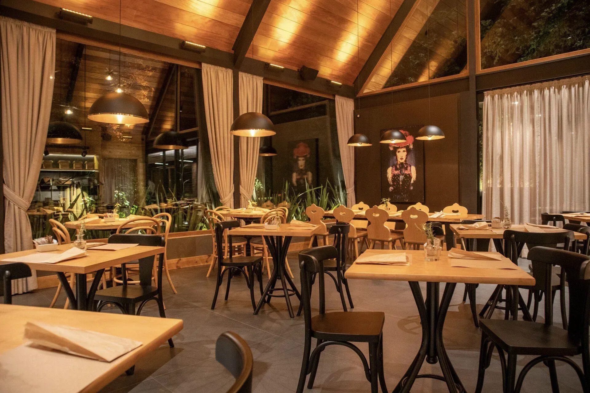 Interior de restaurante aconchegante e acolhedor, com mesas de madeira, cadeiras pretas, luminárias pendentes e cortinas altas.
