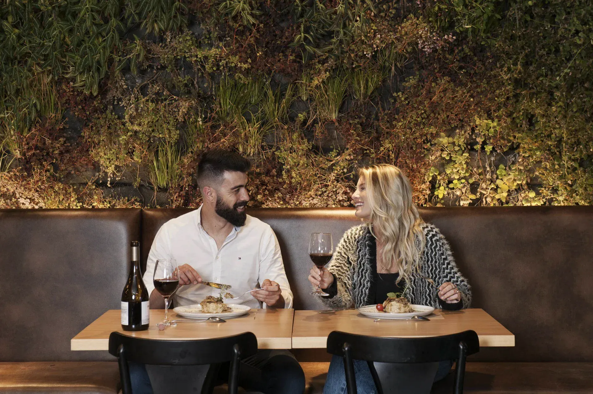 Duas pessoas jantando em uma mesa de restaurante, conversando enquanto bebem e comem em um ambiente aconchegante e iluminado.