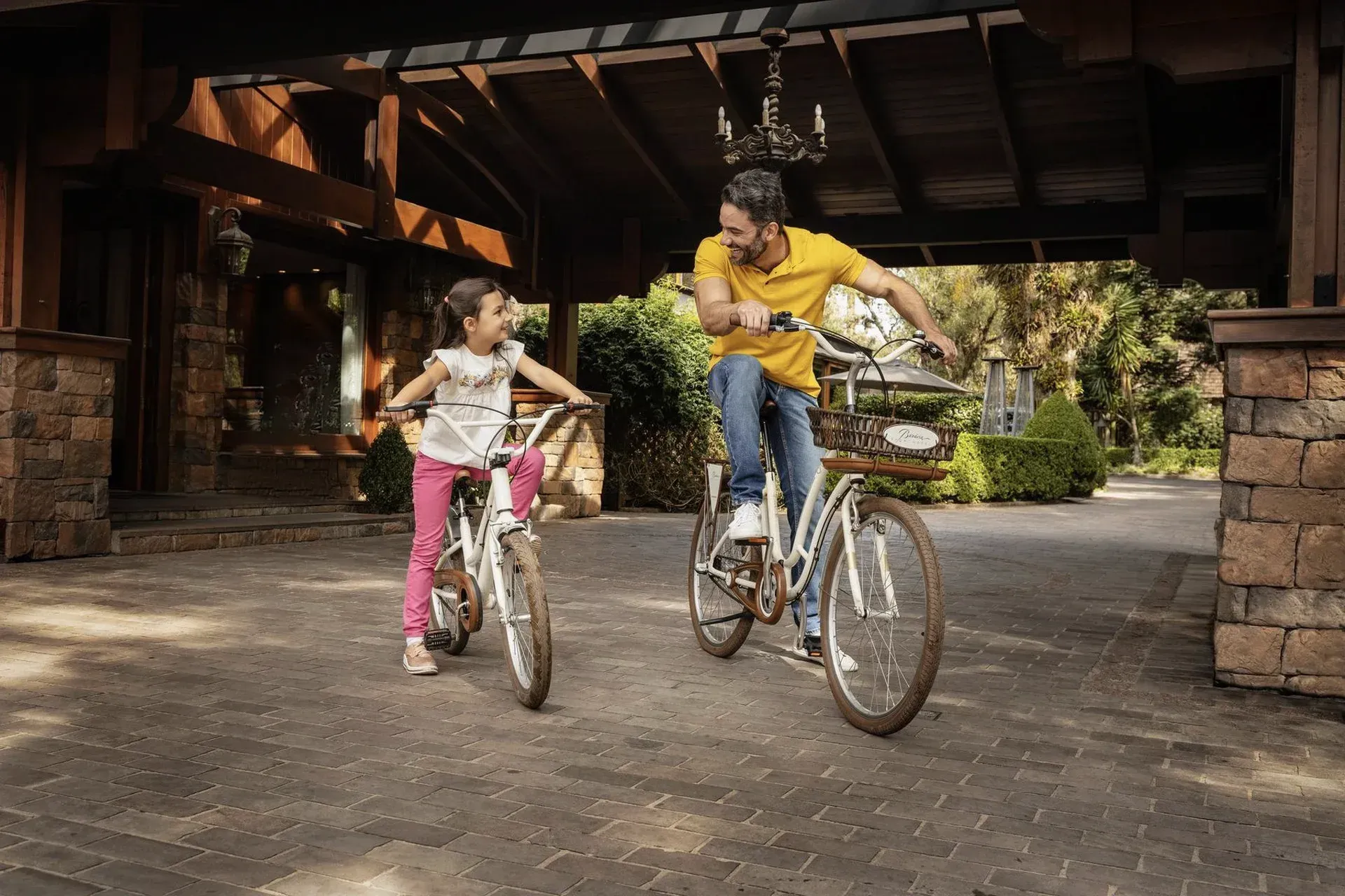 Adulto e criança andando de bicicleta sob um abrigo de madeira em um pátio rústico.