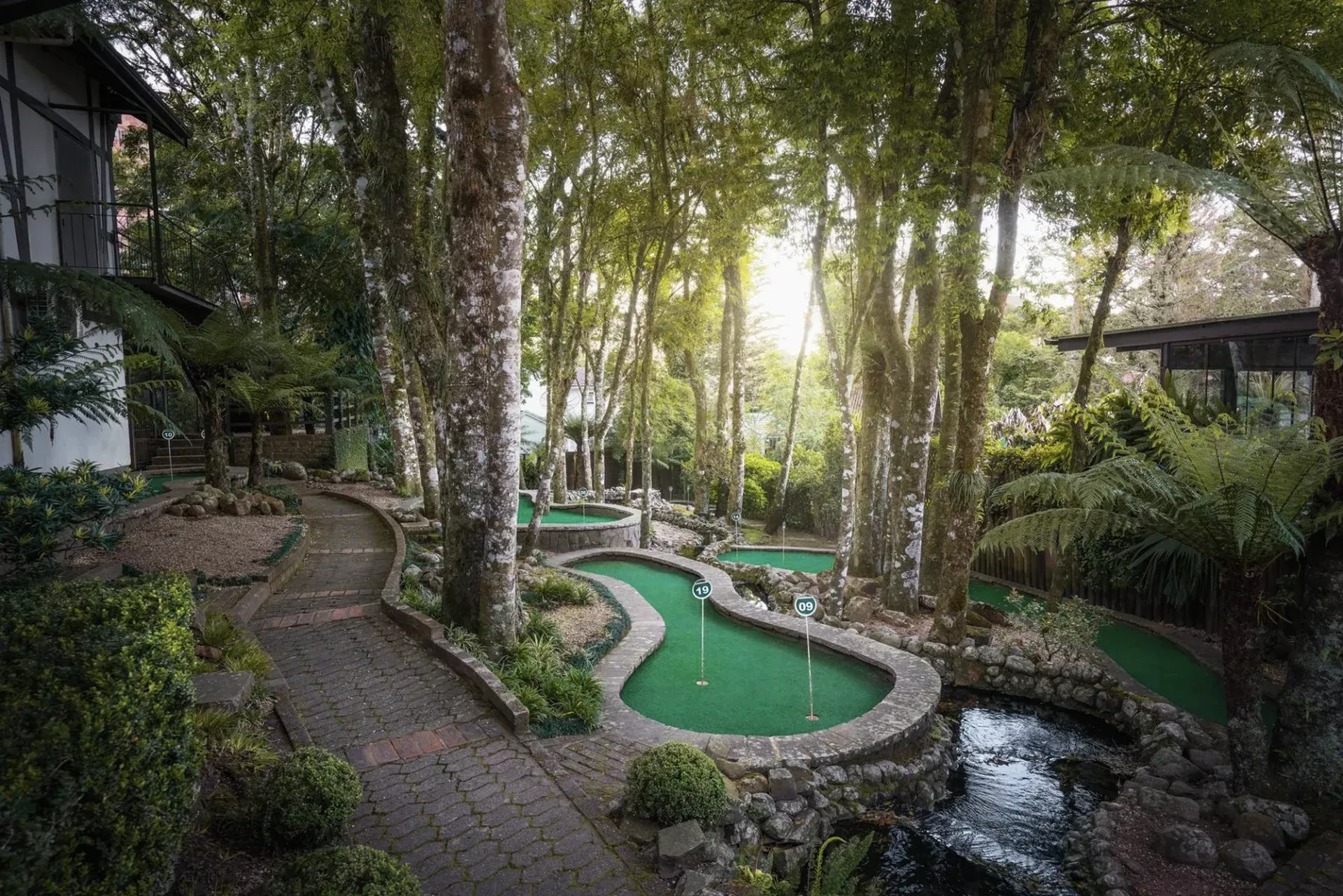 Jardim arborizado com caminhos sinuosos de pedra, lagos verdes e cabanas de palha sombreadas.