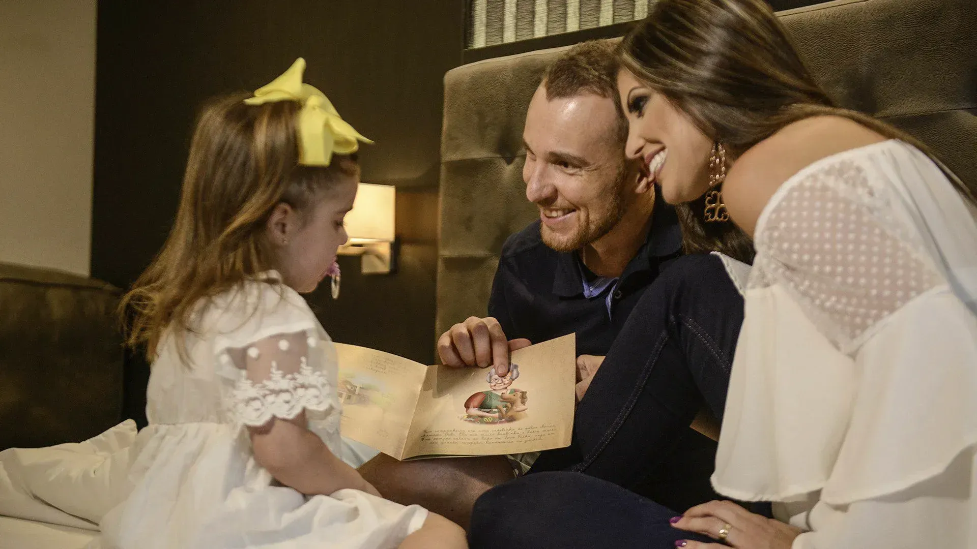 Família lendo um livro de histórias junta na cama, sorrindo em um quarto aconchegante e acolhedor.