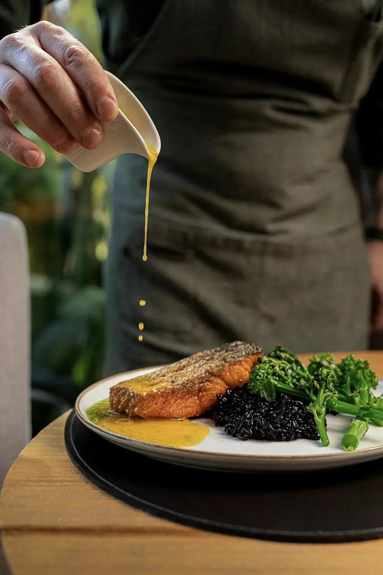 Chef regando o molho sobre um prato de salmão com verduras e arroz negro.