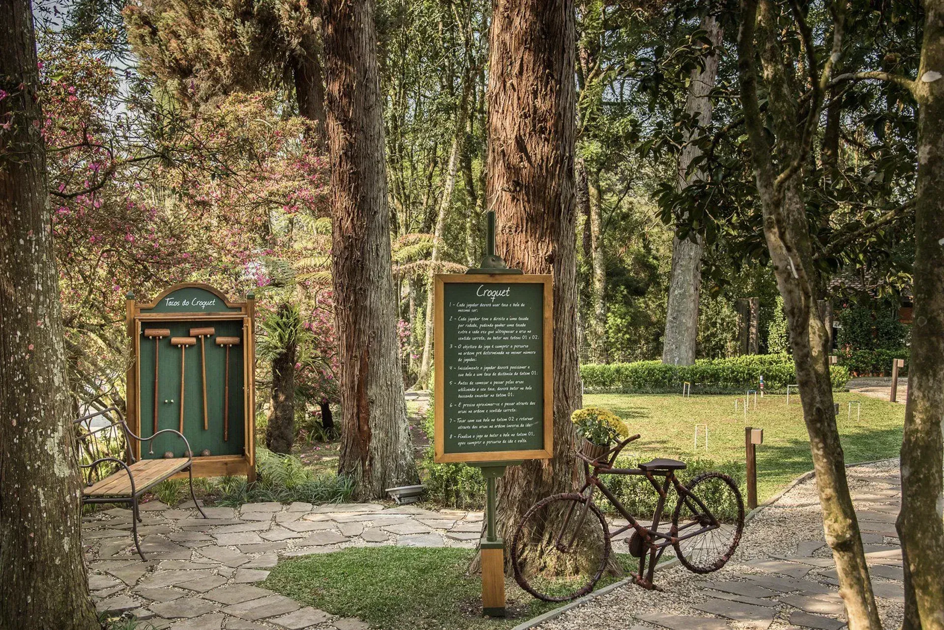 Entrada do parque com árvores, uma placa verde e uma bicicleta ao lado de um caminho de paralelepípedos.