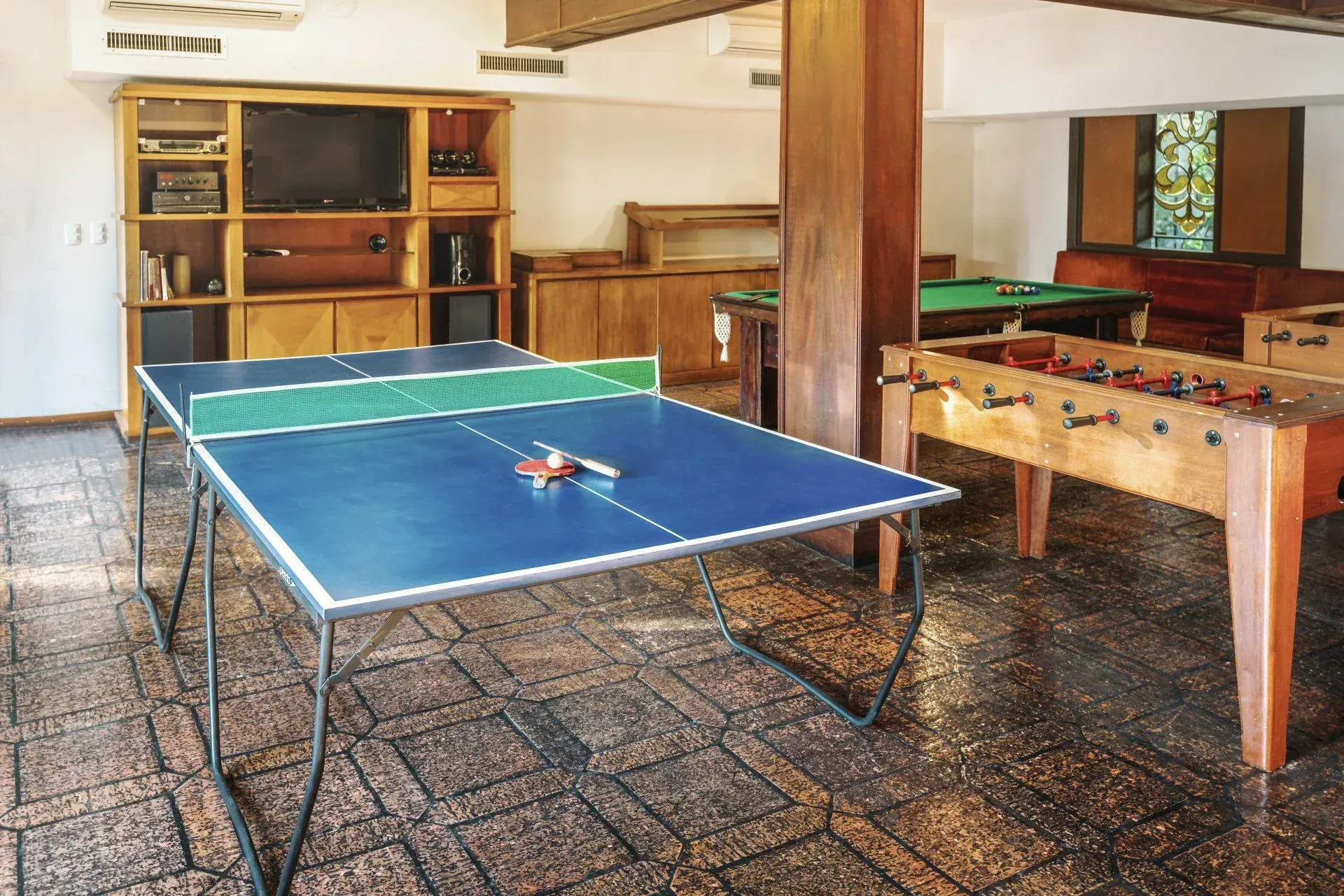 Mesa de pingue-pongue azul em uma sala de jogos, ao lado de uma mesa de pebolim e prateleiras de madeira.