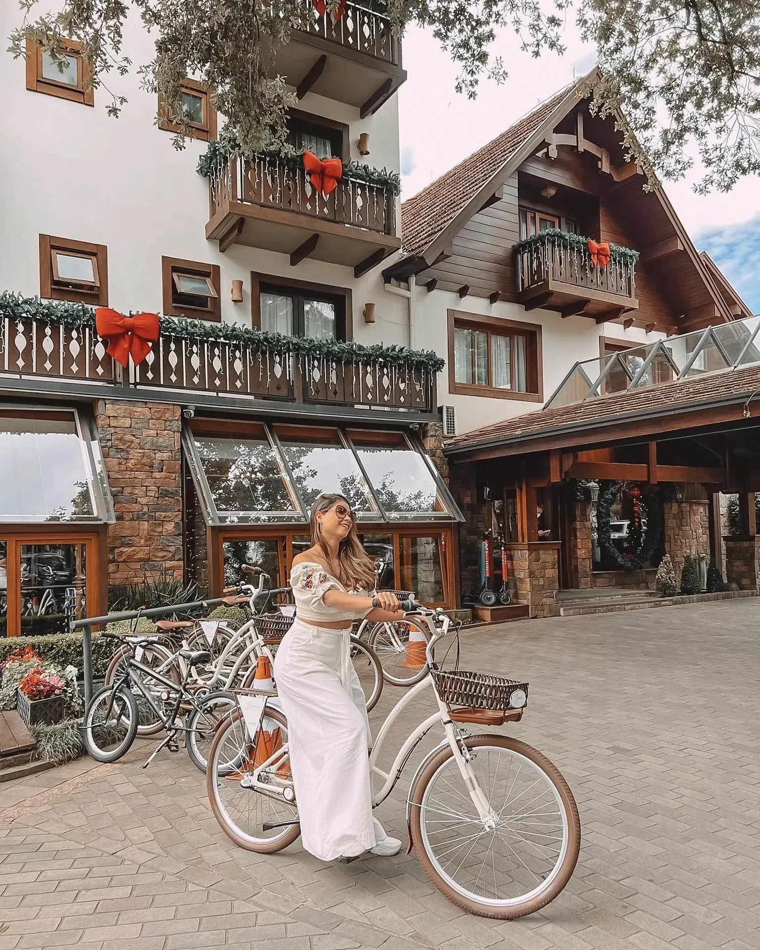 Mulher de vestido branco posando com uma bicicleta em frente a um edifício rústico em estilo alpino.
