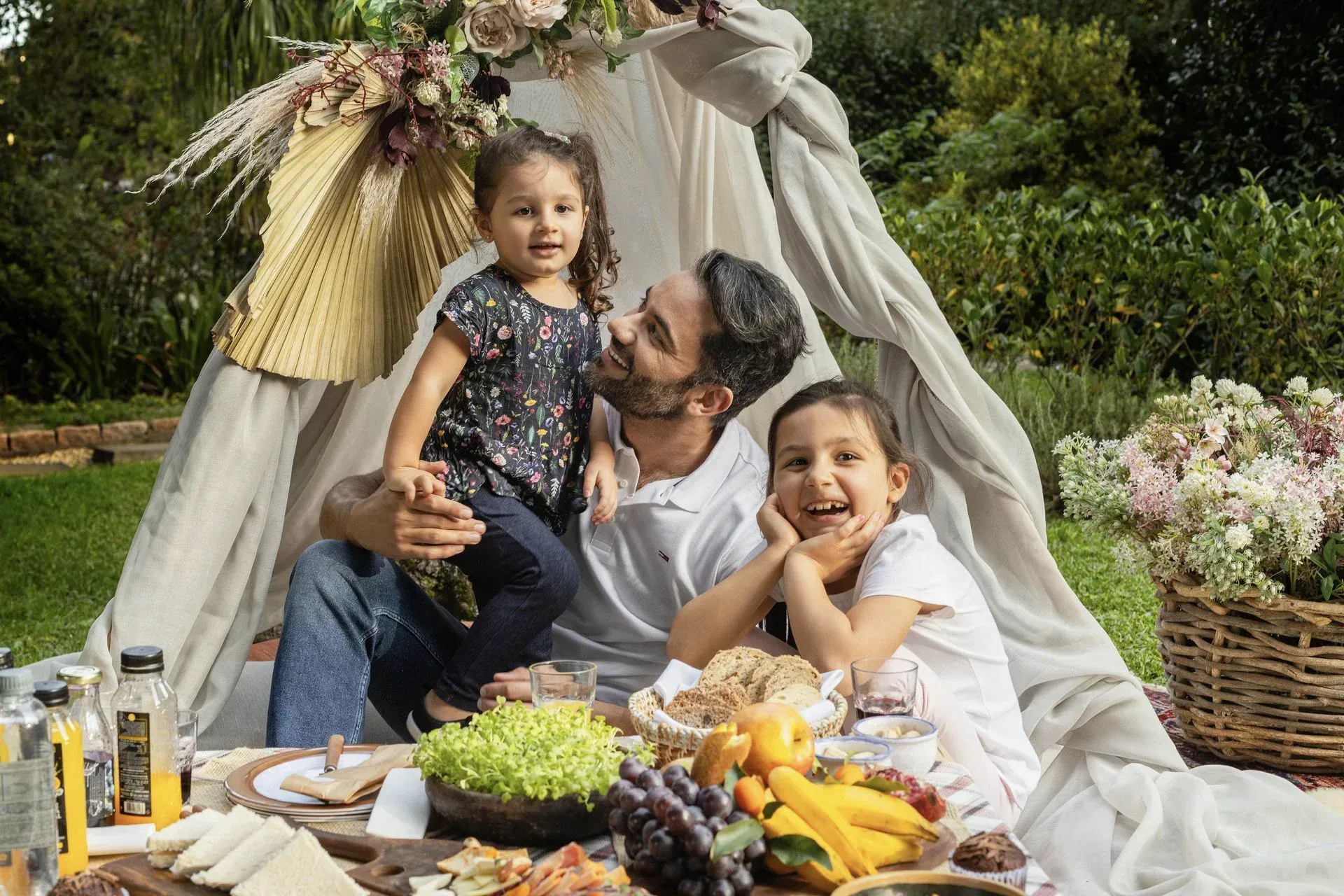 Família fazendo piquenique em uma manta em um jardim com frutas, flores e uma pequena tenda ao fundo.