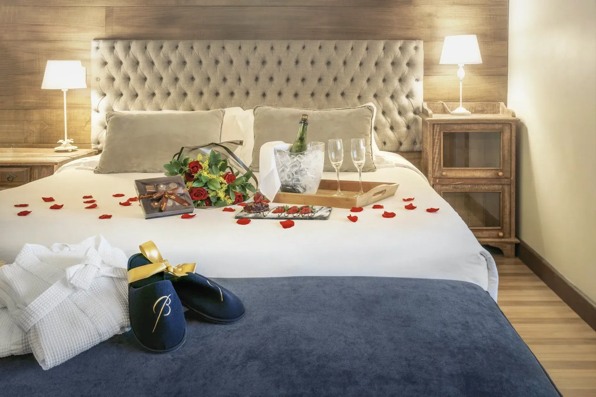 Cama de hotel romântica com pétalas de rosa, champanhe, flores e dois abajures nas mesas de cabeceira.