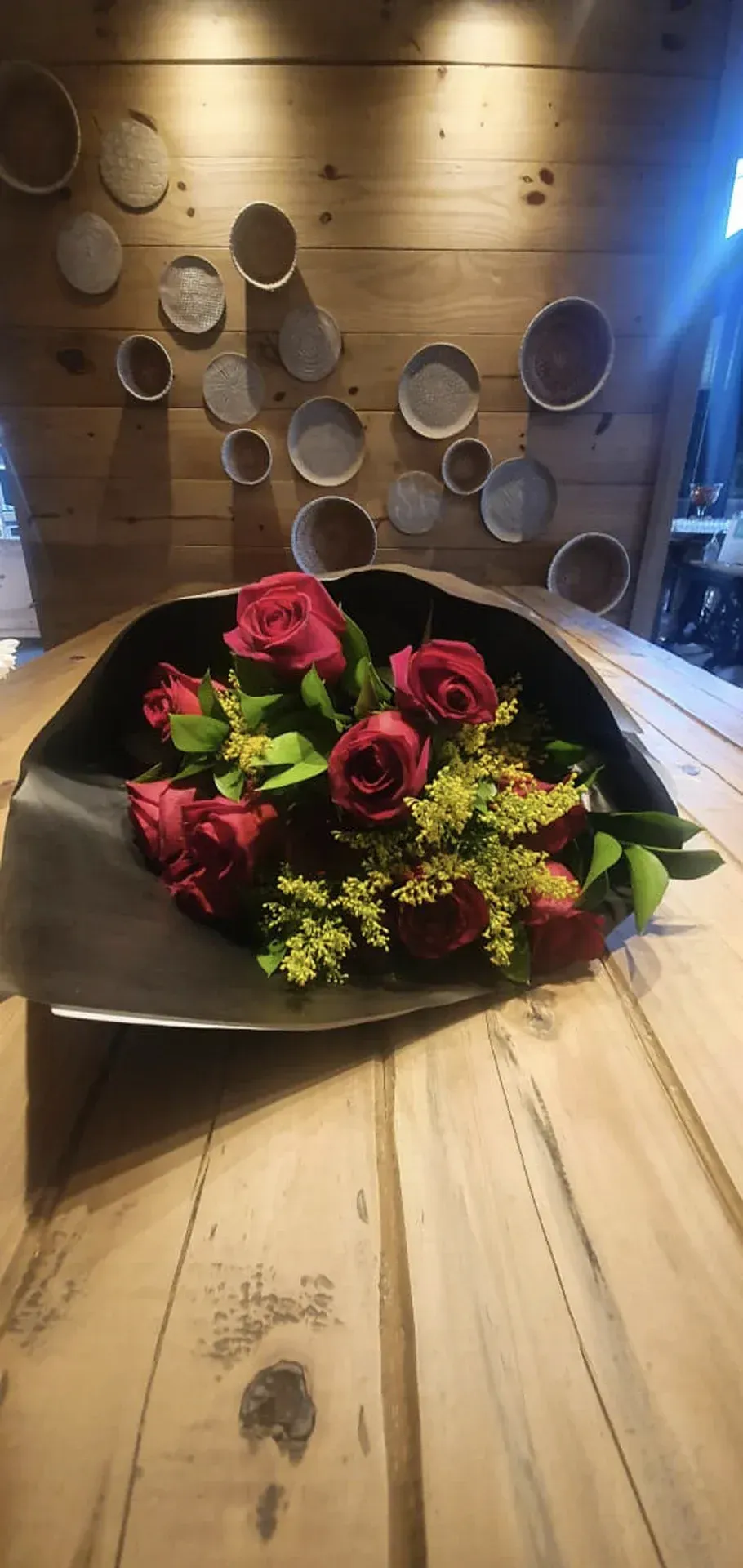 Rosas vermelhas e flores verdes num vaso preto sobre uma mesa de madeira.