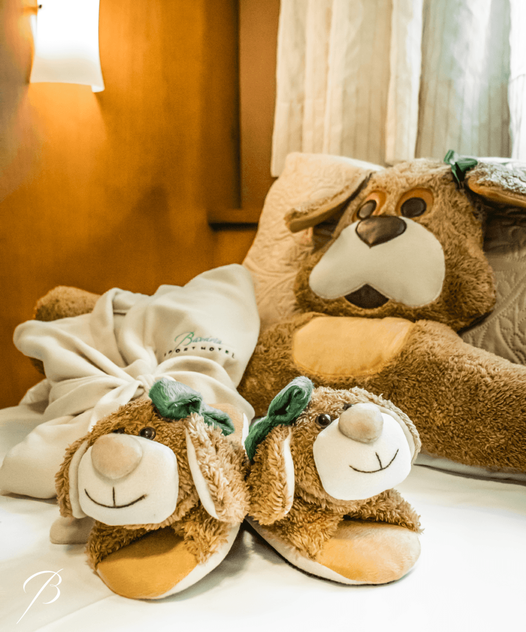 Pantufas de pelúcia em formato de gato e uma almofada de gato em uma cama perto de uma janela com luz quente.
