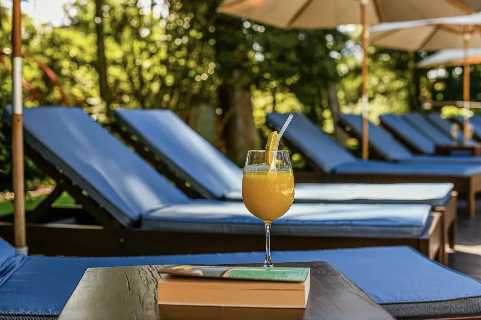 Espreguiçadeiras azuis com guarda-sóis ao lado de uma mesa com uma bebida e um livro junto à piscina.