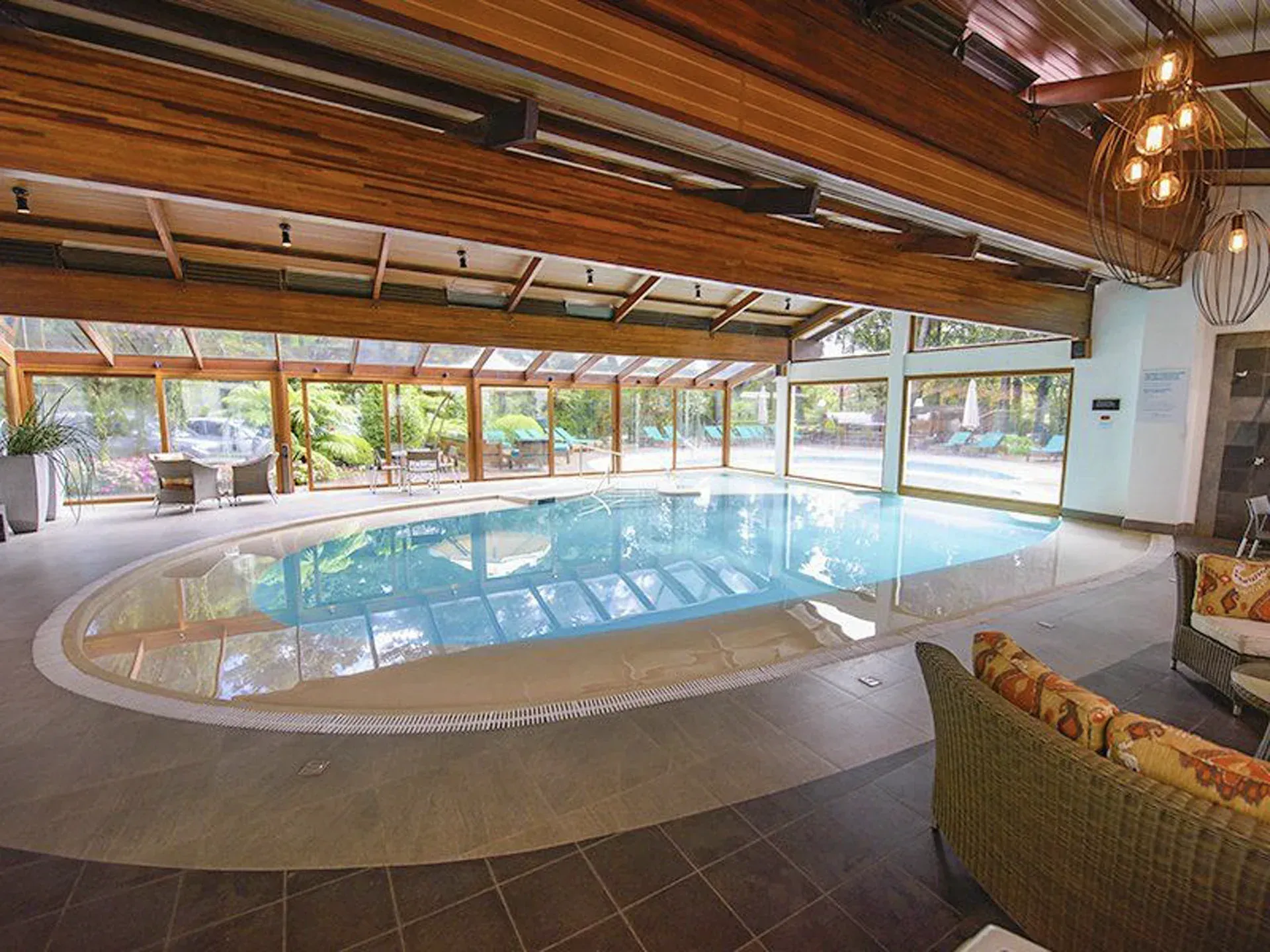 Piscina coberta de resort com lagoa azul curva, teto com vigas de madeira e espreguiçadeiras junto a grandes janelas.