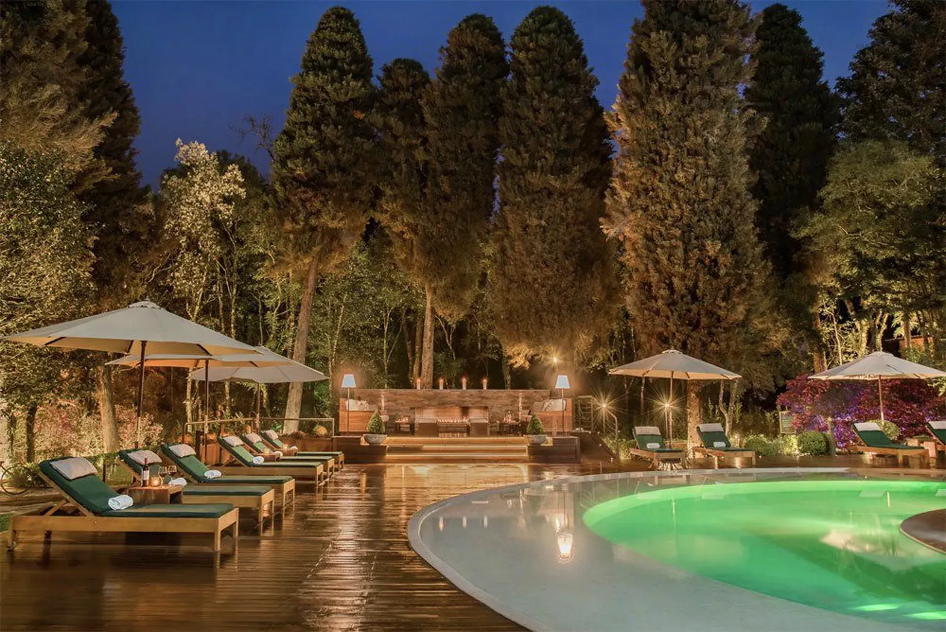 Resort noturno à beira da piscina com espreguiçadeiras, guarda-sóis e piscina verde fluorescente ao lado de árvores.