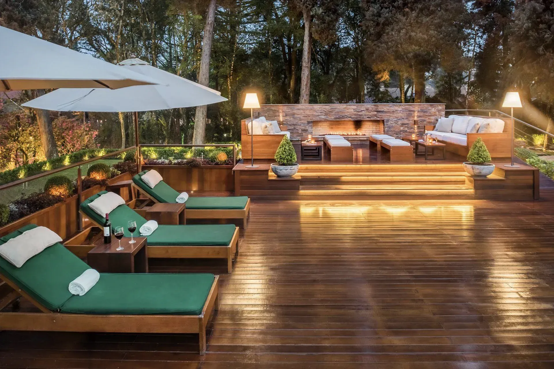 Terraço ao ar livre ao entardecer com espreguiçadeiras verdes, guarda-sóis brancos e assentos à luz de velas junto à água.