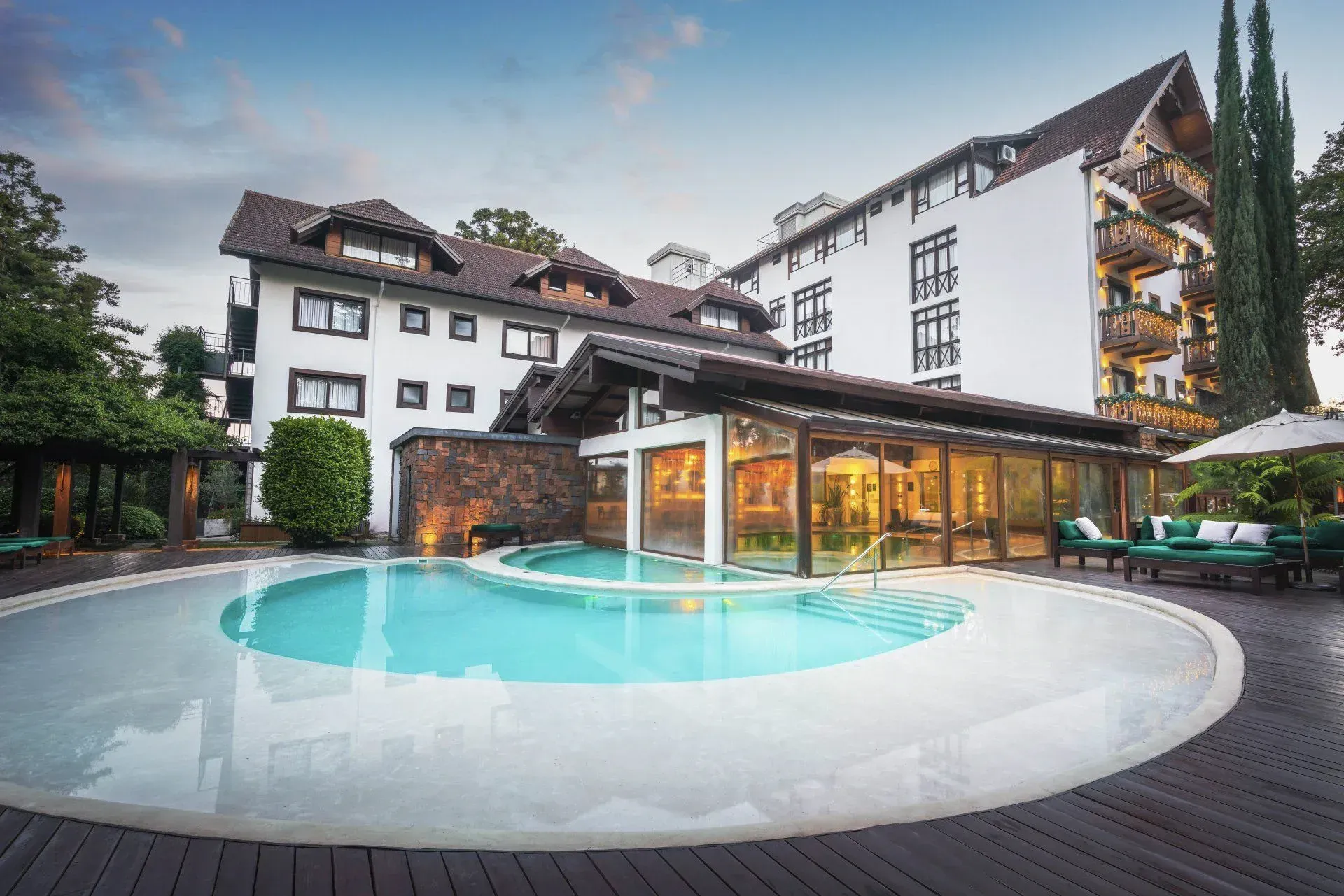 Pátio de hotel de luxo com piscina curva e edifícios brancos ao entardecer.