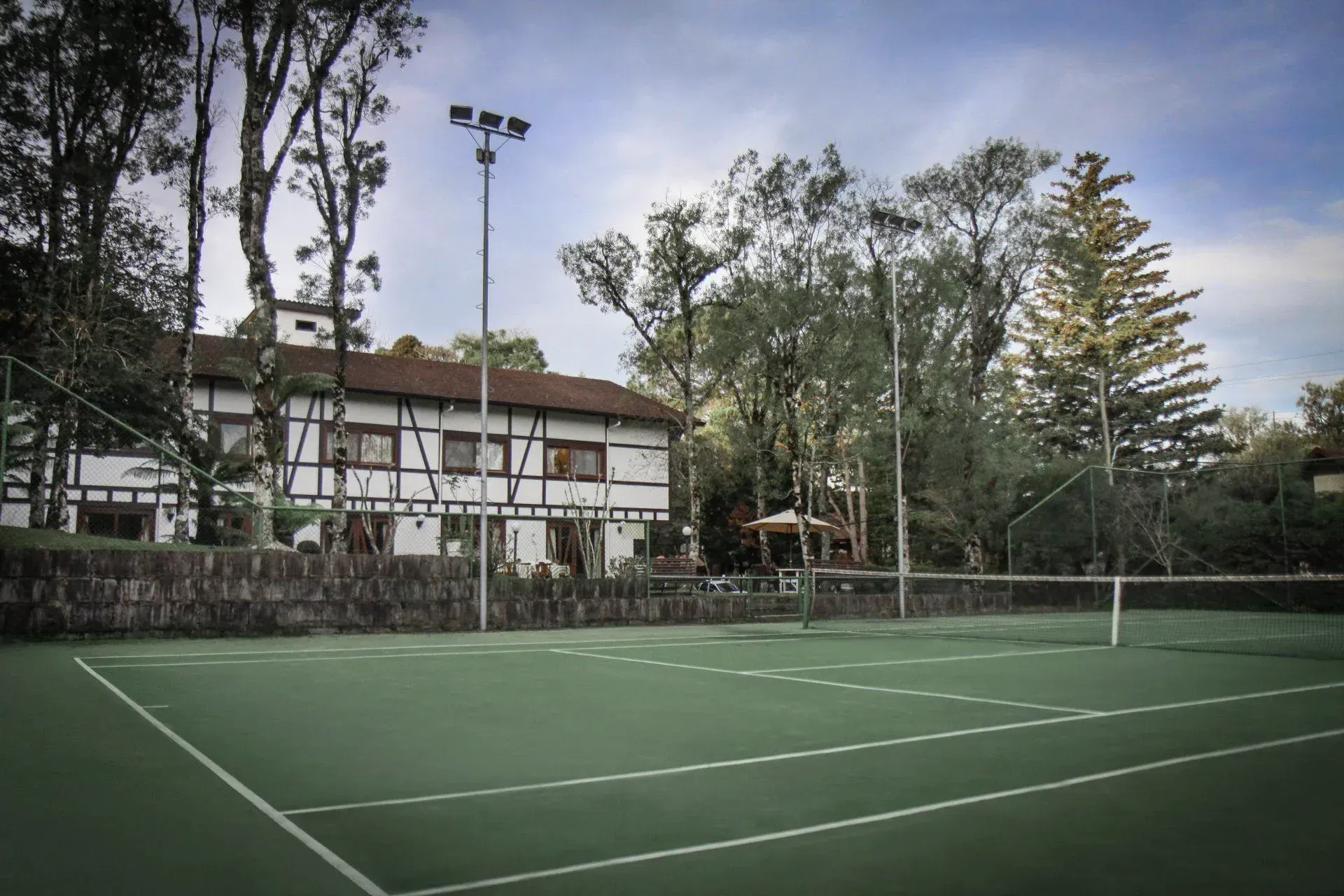 Quadra de tênis vazia ao lado de um clube branco e árvores altas ao entardecer.