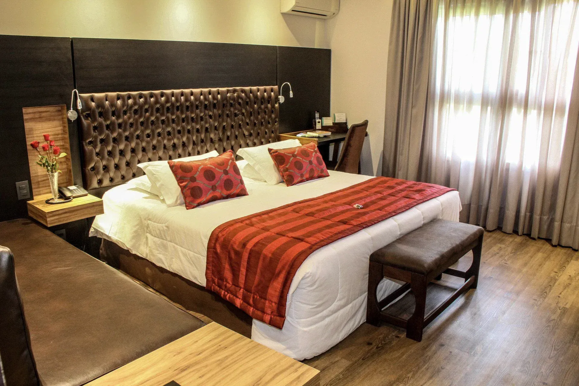 Quarto de hotel com cama arrumada, caminho de mesa vermelho, cabeceira estofada e grande janela com cortina.