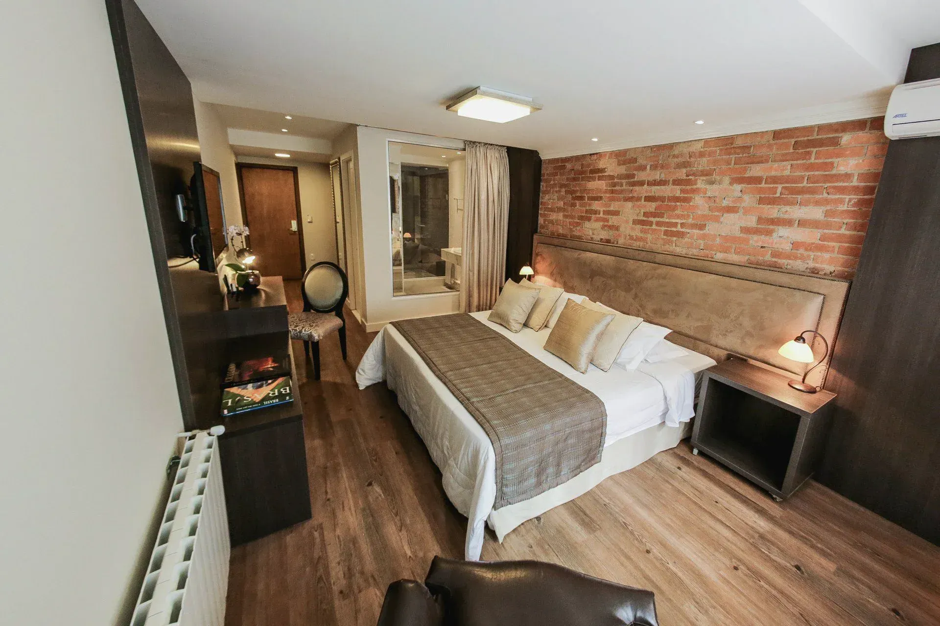 Quarto aconchegante com parede de tijolos à vista, cama de casal, piso de madeira e abajures.