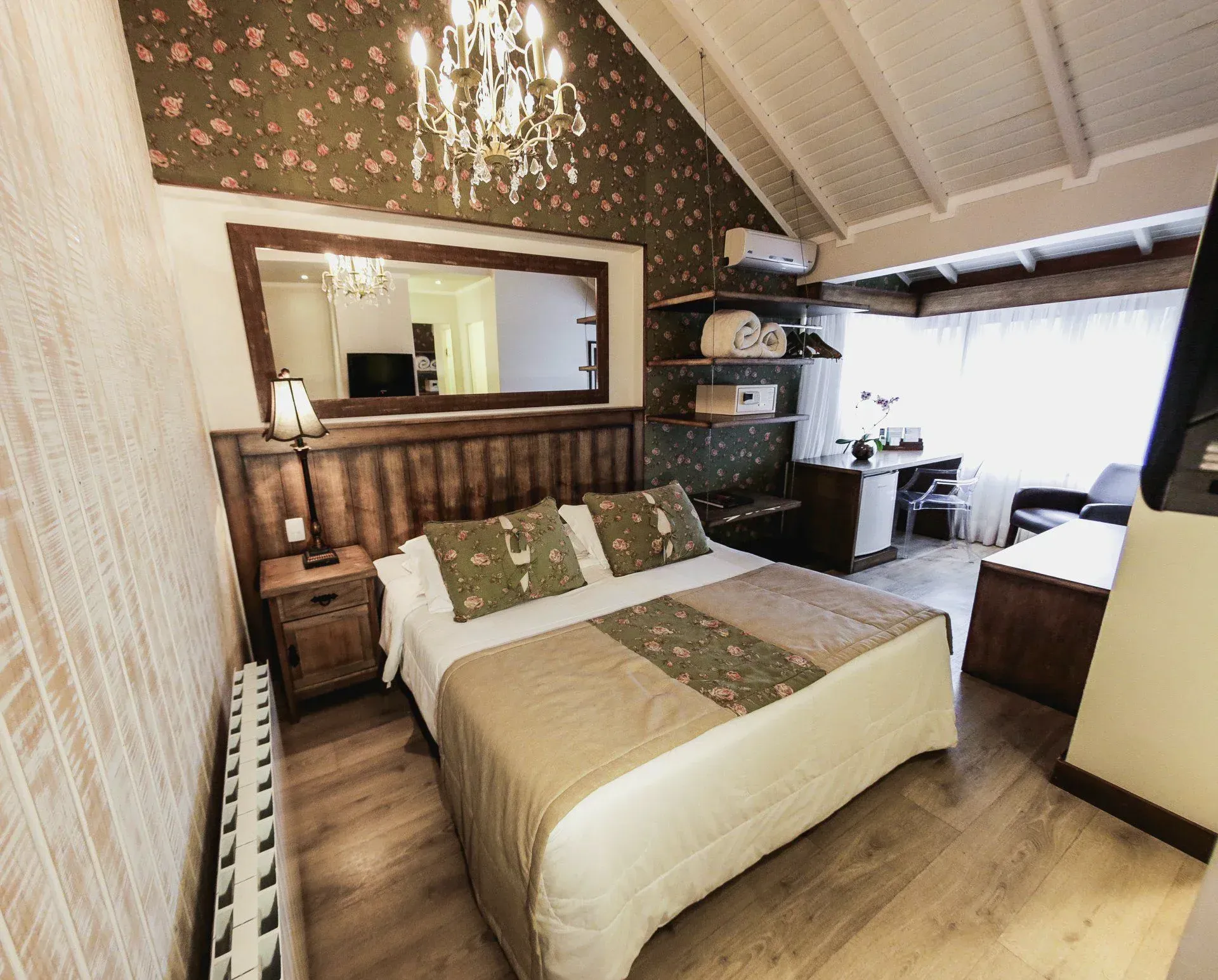 Quarto aconchegante com cama, cabeceira espelhada, papel de parede estampado e uma área de estar com janela.