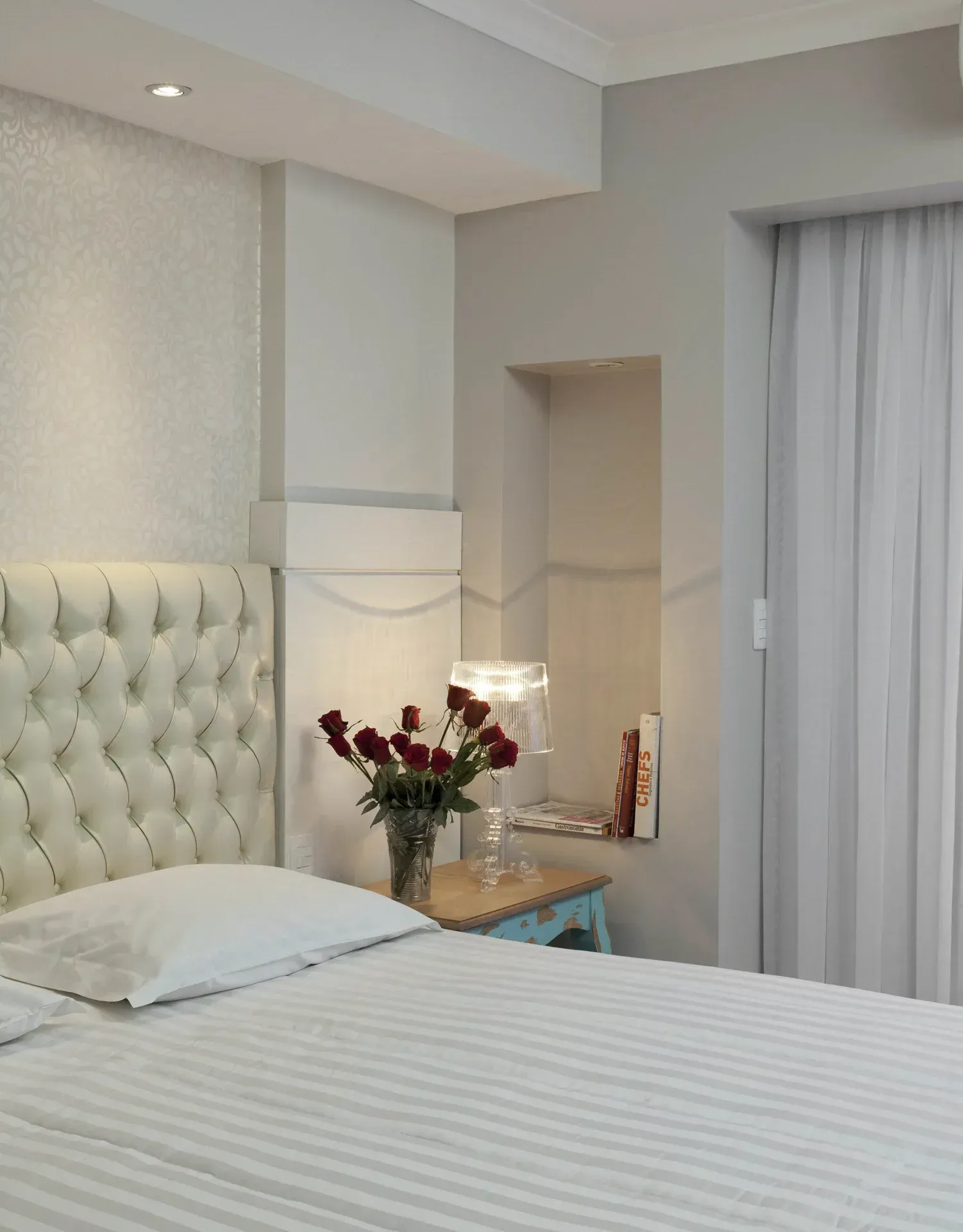 Quarto elegante com colcha listrada, cabeceira estofada, flores na mesa de cabeceira e cortinas transparentes.