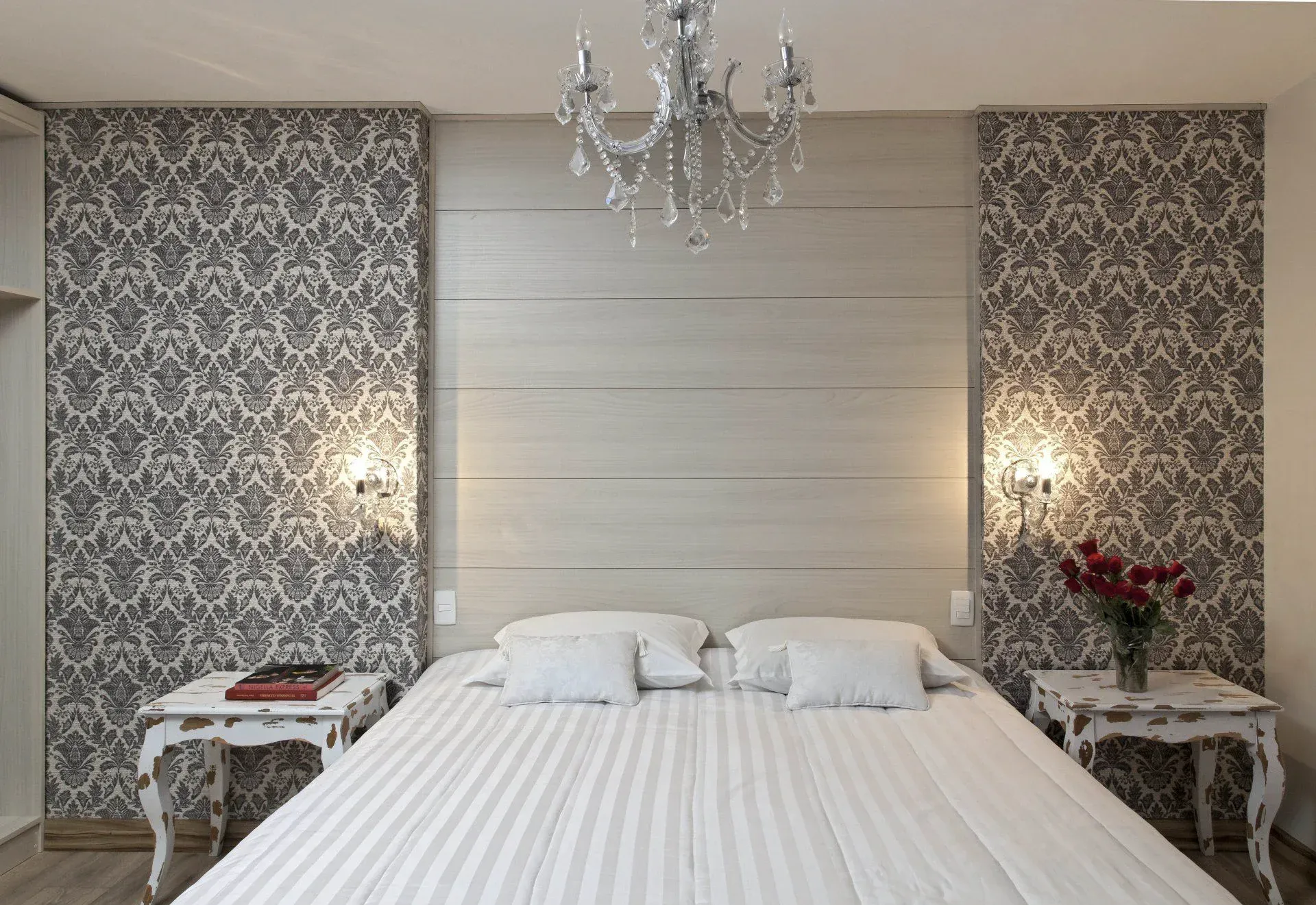 Quarto elegante com uma grande cama branca, papel de parede estampado, lustre e mesas de cabeceira com abajures.