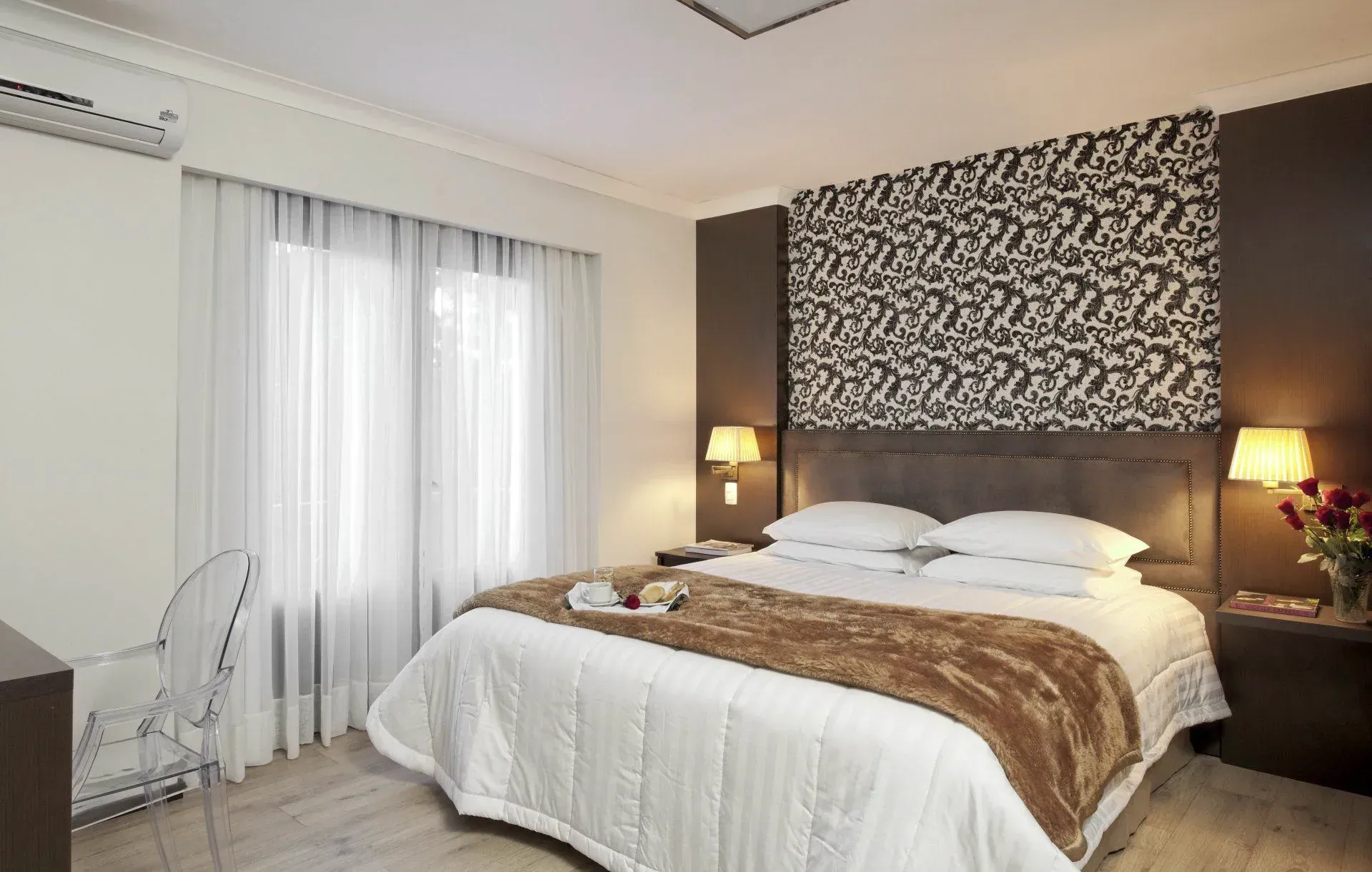 Quarto luminoso com uma cama grande, cabeceira estampada, cortinas brancas e abajures com luz quente.