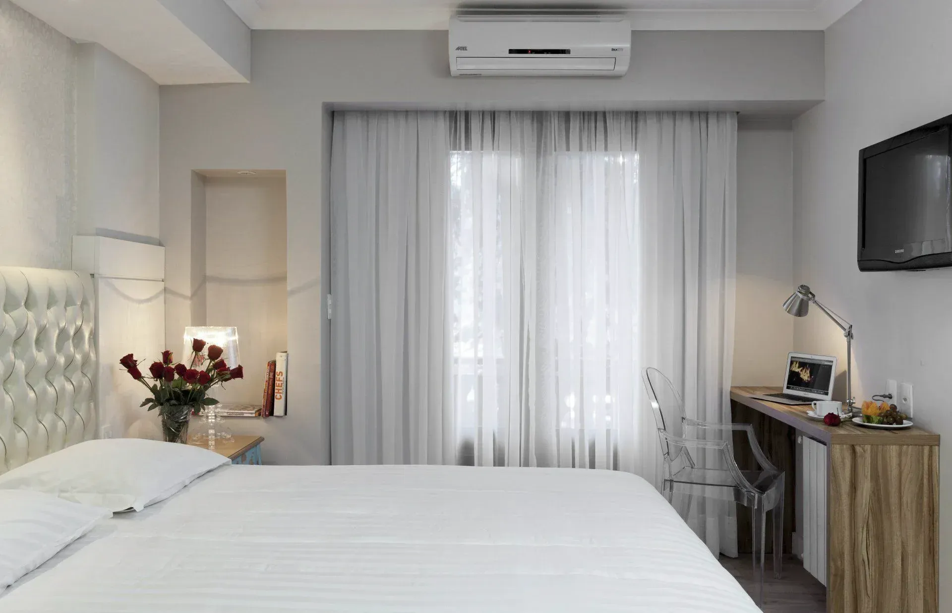 Quarto de hotel impecável com cama branca, cortinas na janela, escrivaninha, TV de parede e flores na mesa de cabeceira.