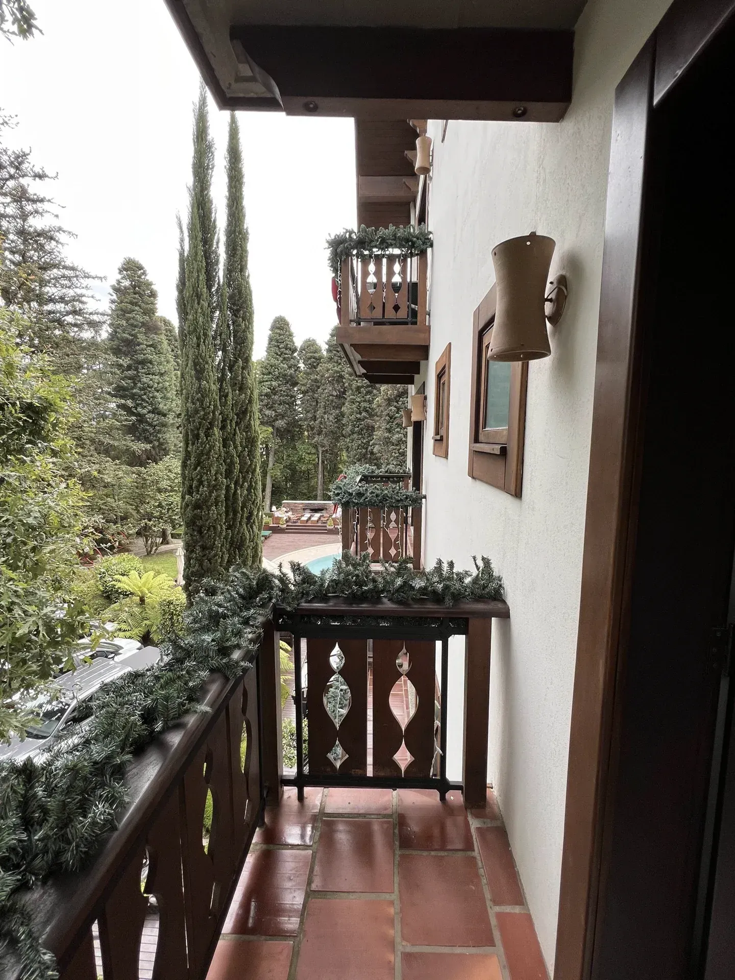 Varanda estreita com piso de ladrilho vermelho, corrimão de madeira, planta suspensa e vista para árvores altas ao lado de um prédio branco.