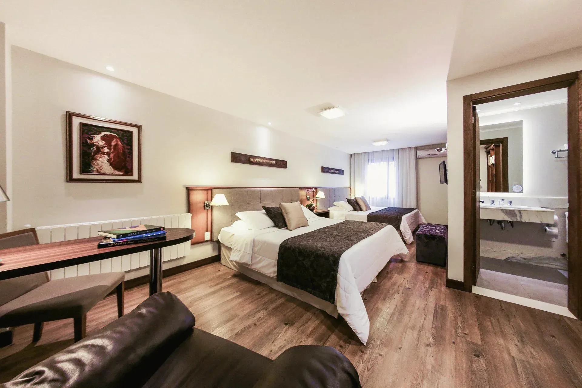 Quarto de hotel moderno com cama arrumada, escrivaninha, poltrona e banheiro visível através de uma porta aberta.