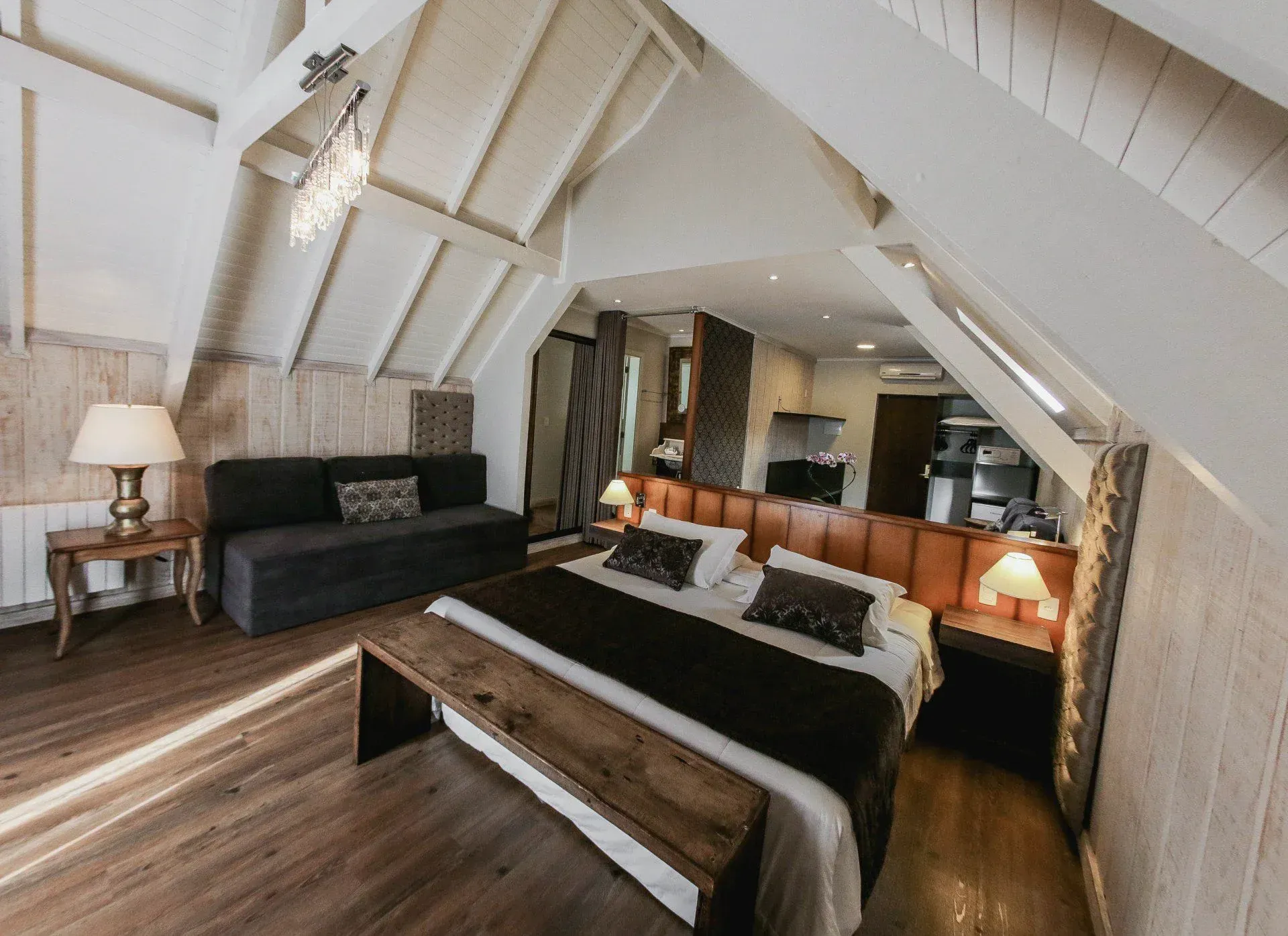 Quarto aconchegante no sótão com paredes brancas inclinadas, sofá escuro e cama com estrutura de madeira e iluminação quente.