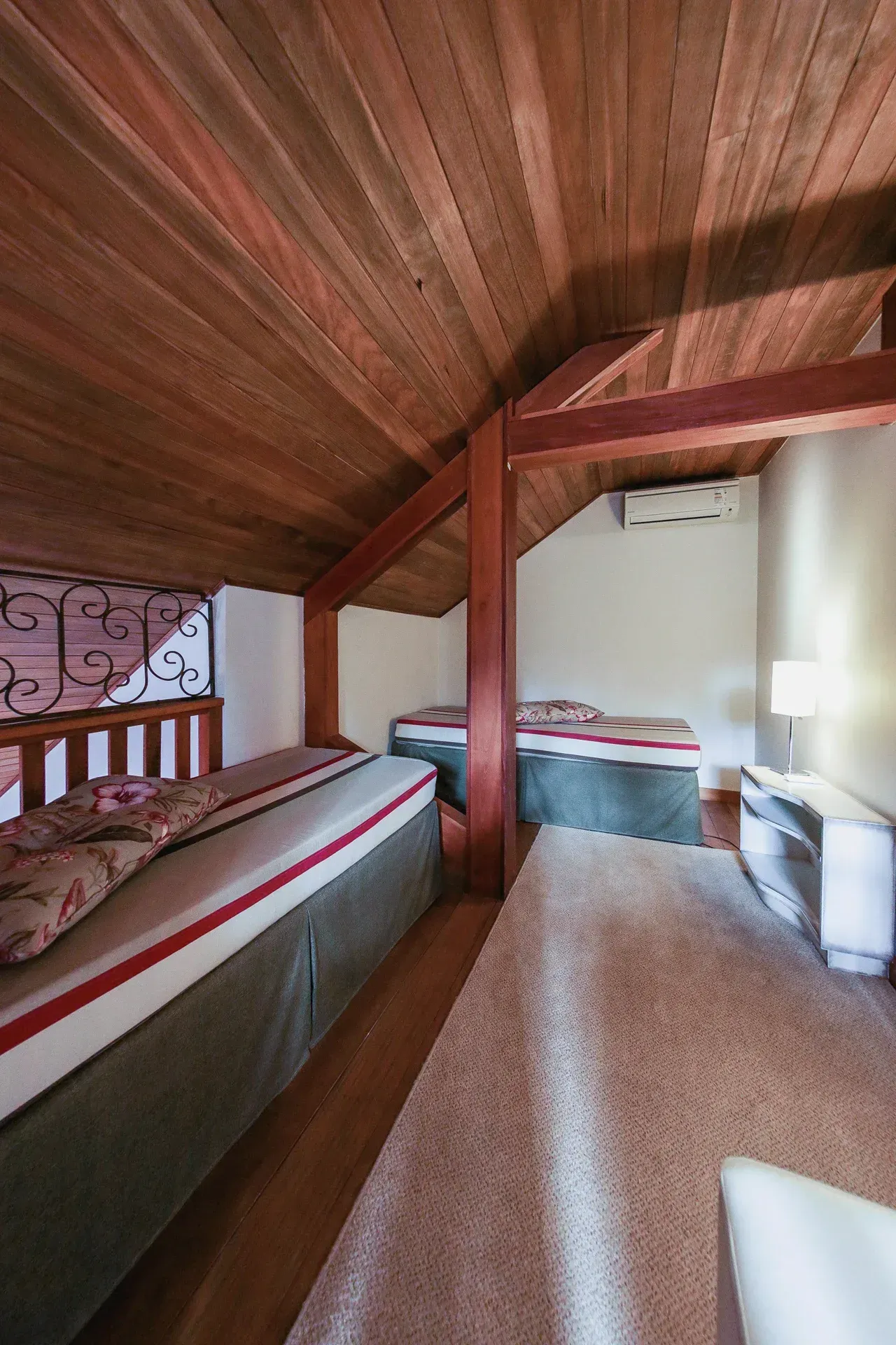 Pequeno quarto no sótão com duas camas de solteiro, teto inclinado com painéis de madeira e um recanto com assento junto à janela.