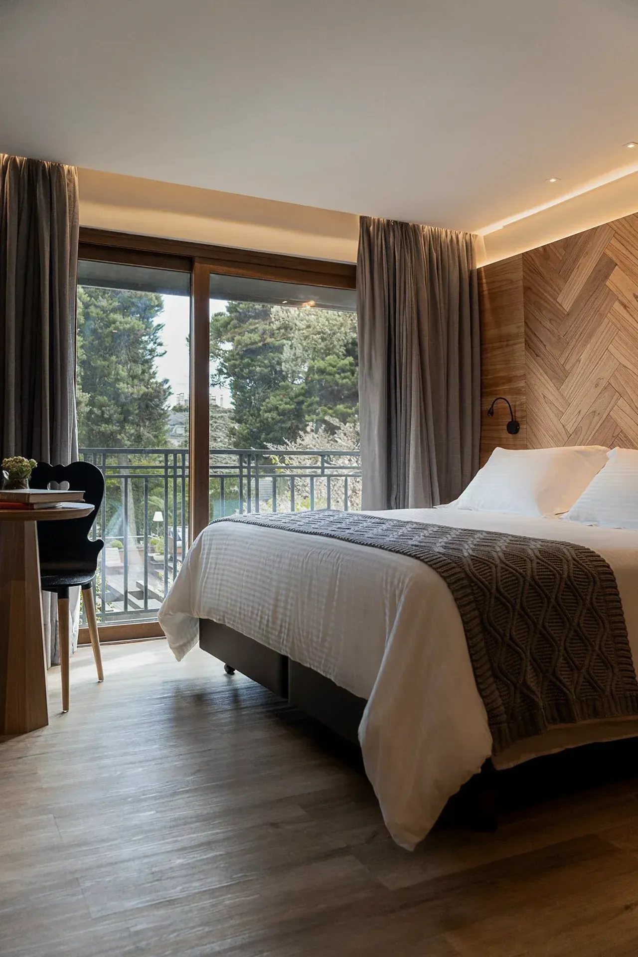 Quarto moderno com cama king-size, janelas do chão ao teto, detalhes em madeira e vista para a varanda.