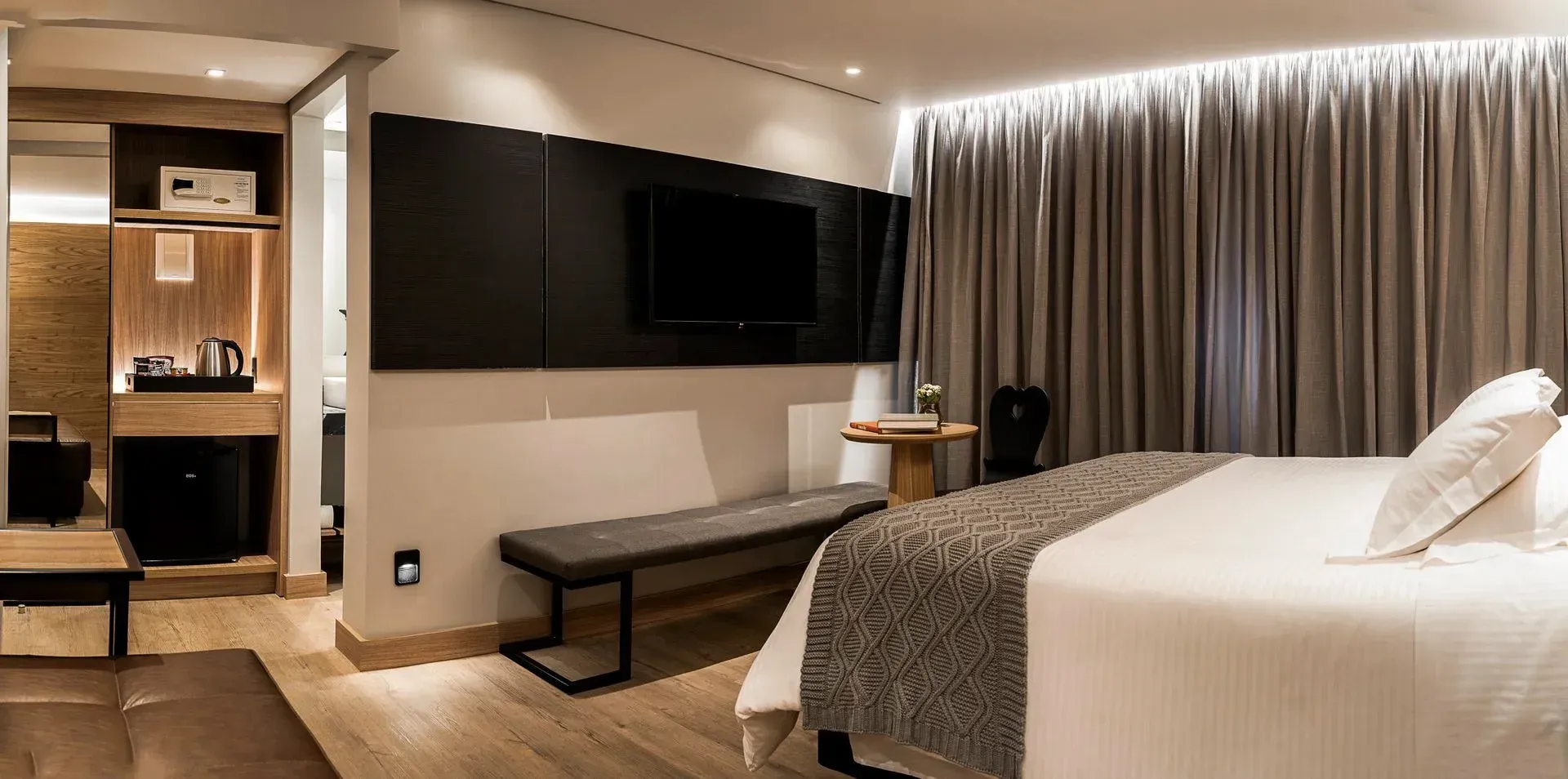 Quarto de hotel moderno com cama grande, TV de parede, escrivaninha e cortinas do chão ao teto.