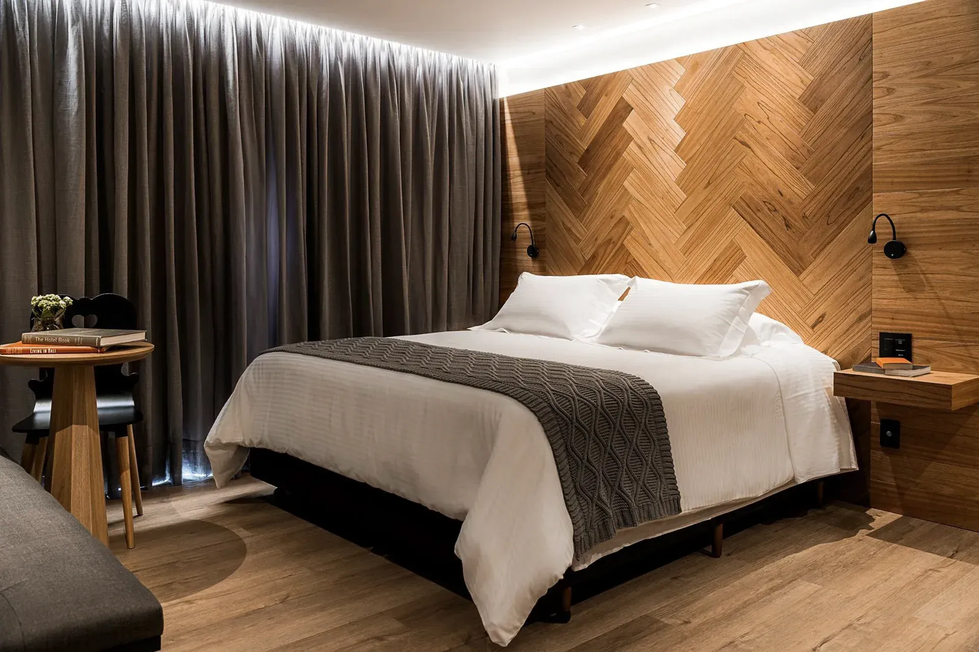Quarto moderno com parede revestida de madeira, cama branca, cortinas escuras e iluminação ambiente aconchegante.