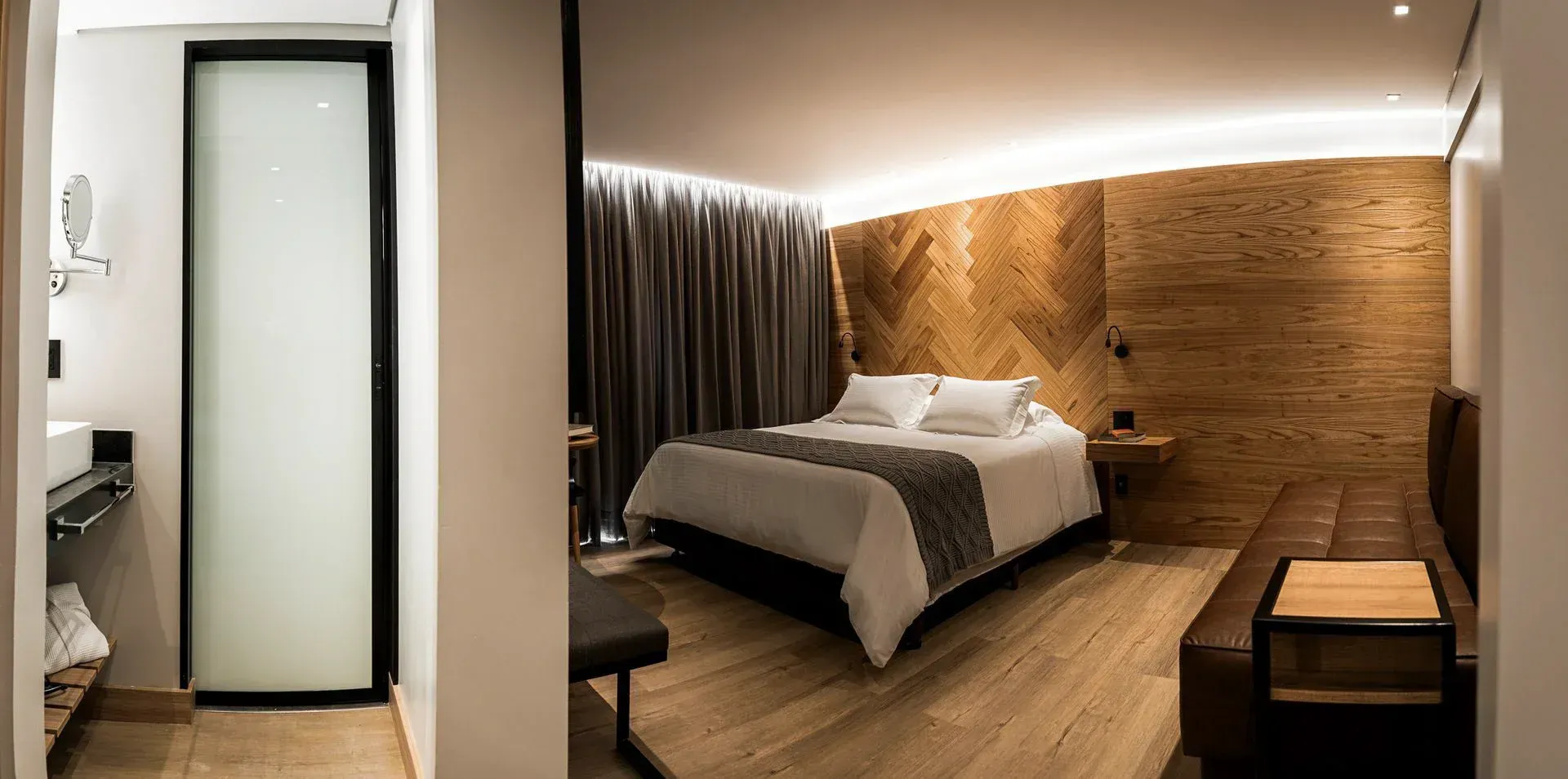 Quarto de hotel moderno com cama, parede de madeira, cortinas e porta do banheiro adjacente visível.
