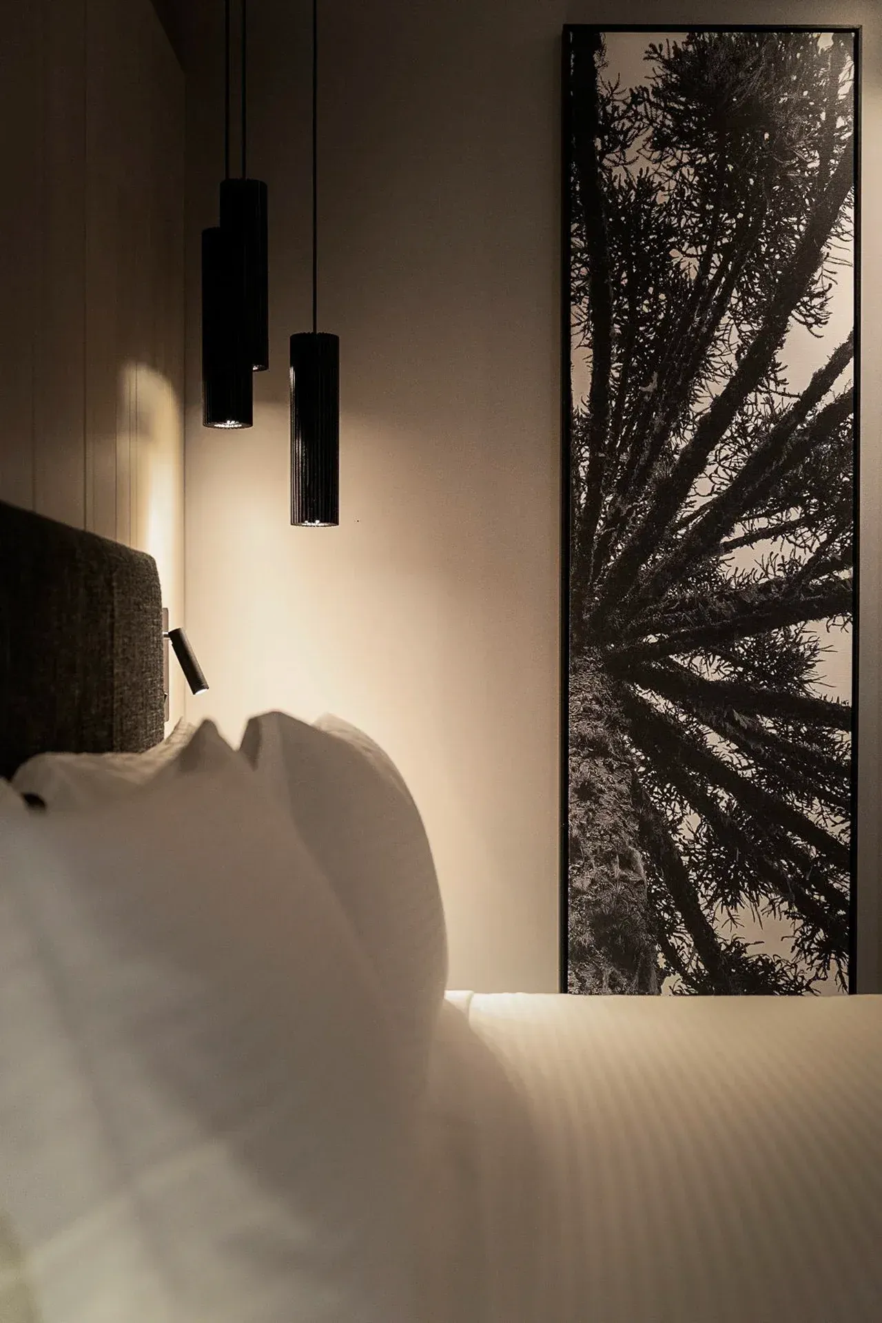 Quarto com pouca luz, roupa de cama branca, iluminação suave de um abajur e vista emoldurada para as árvores pela janela.