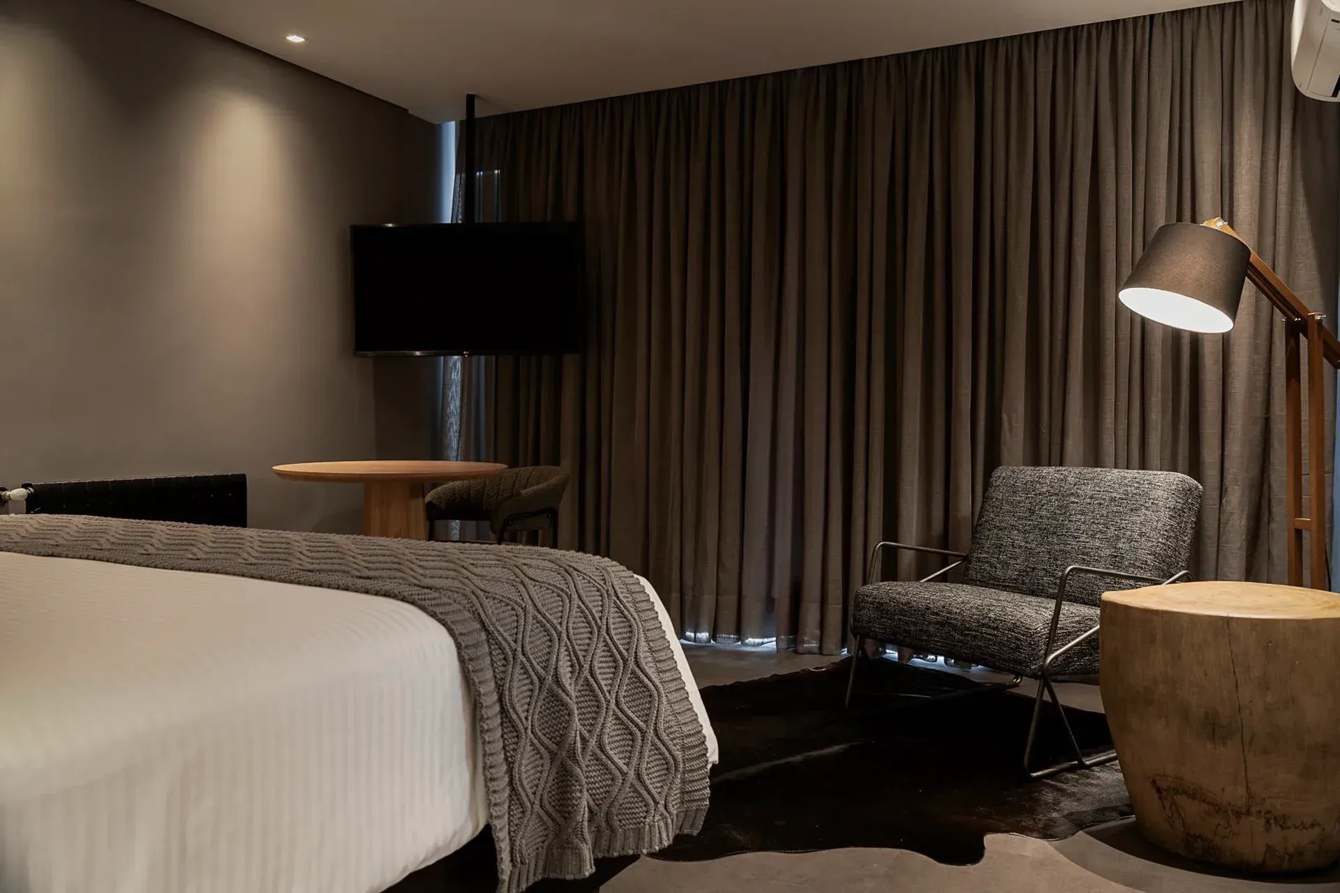 Quarto de hotel moderno com cama, cadeira, luminária de chão, TV e cortinas do chão ao teto.