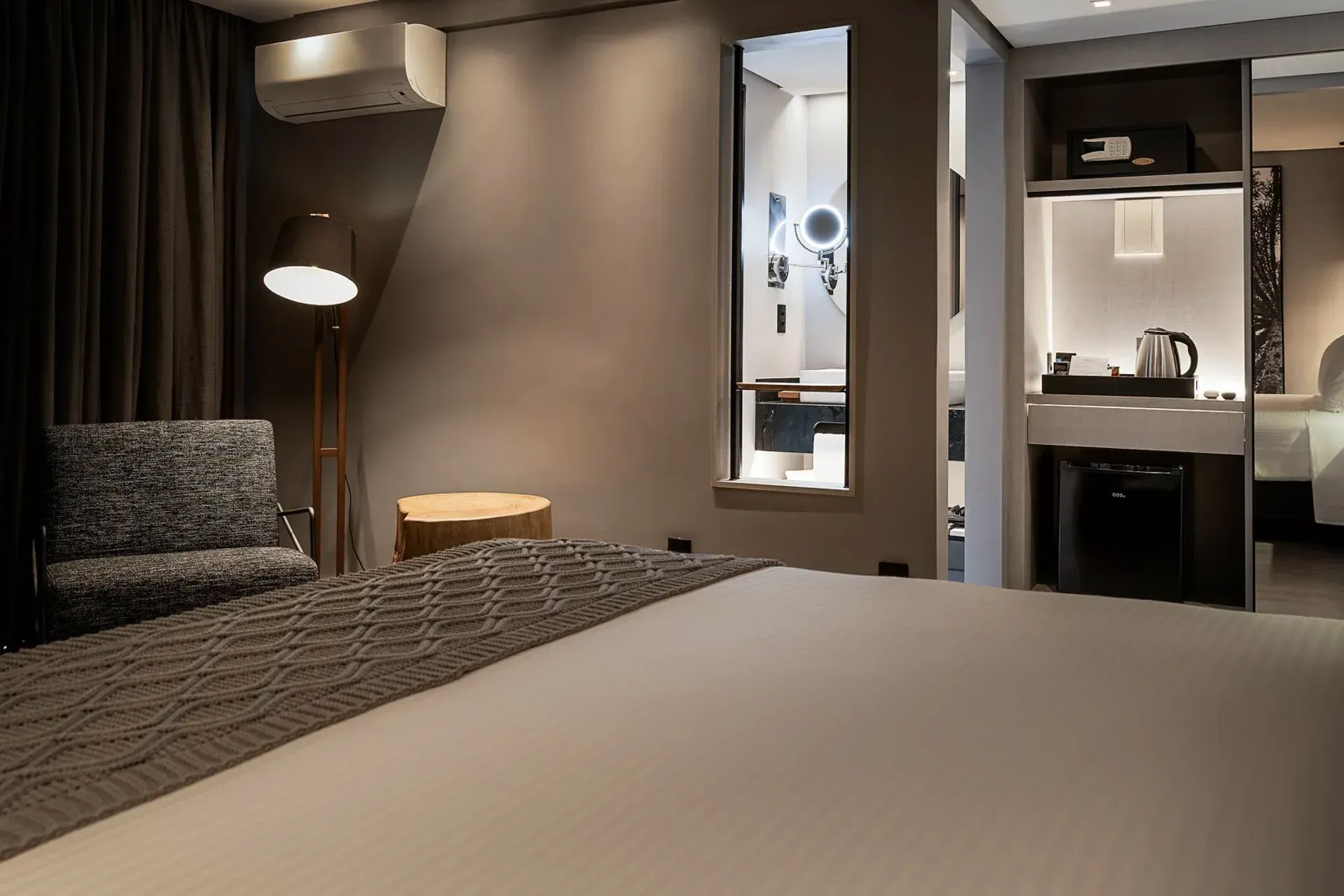 Quarto de hotel moderno com cama grande, luminária de leitura, janela e cozinha compacta.