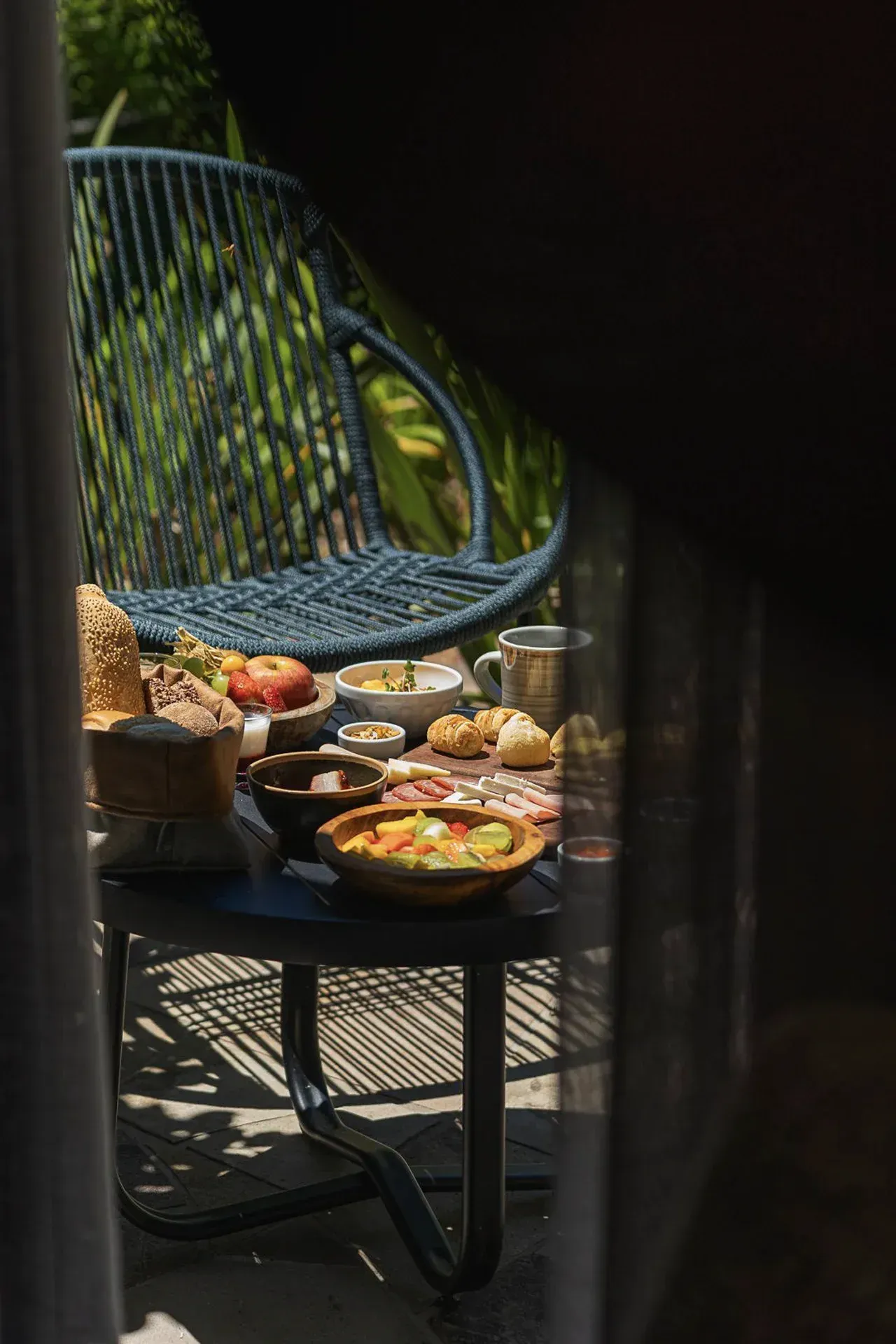 Mesa externa com diversos pratos de café da manhã em um pátio, vista através da estrutura de uma cadeira.